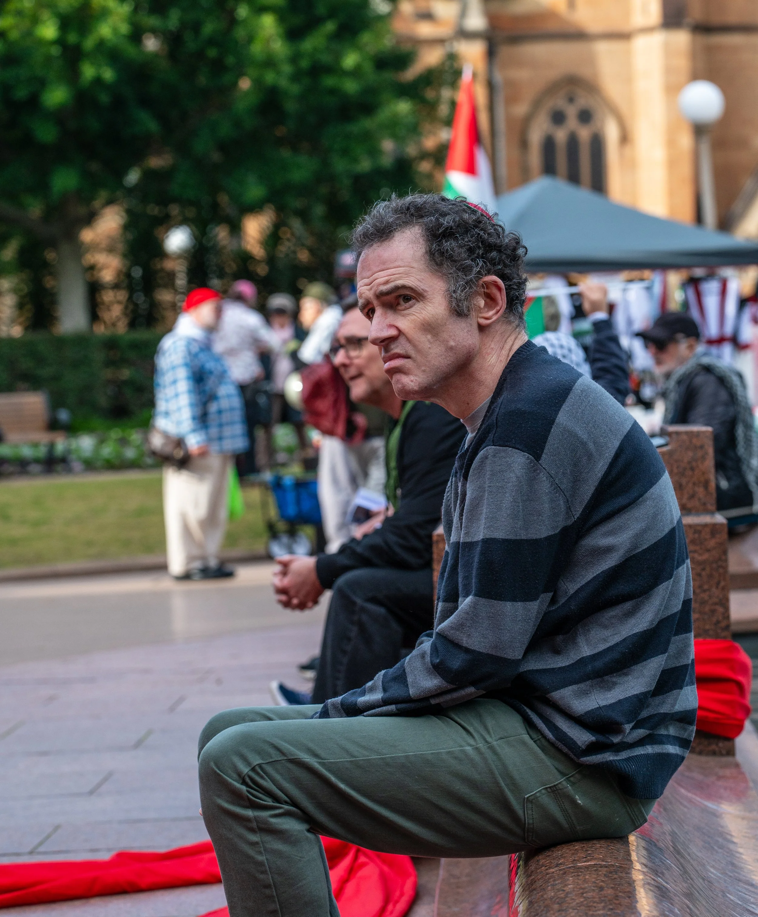 A man sitting on a bench with a serious expression, surrounded by other people at an outdoor event with flags and tents, in front of a large building.