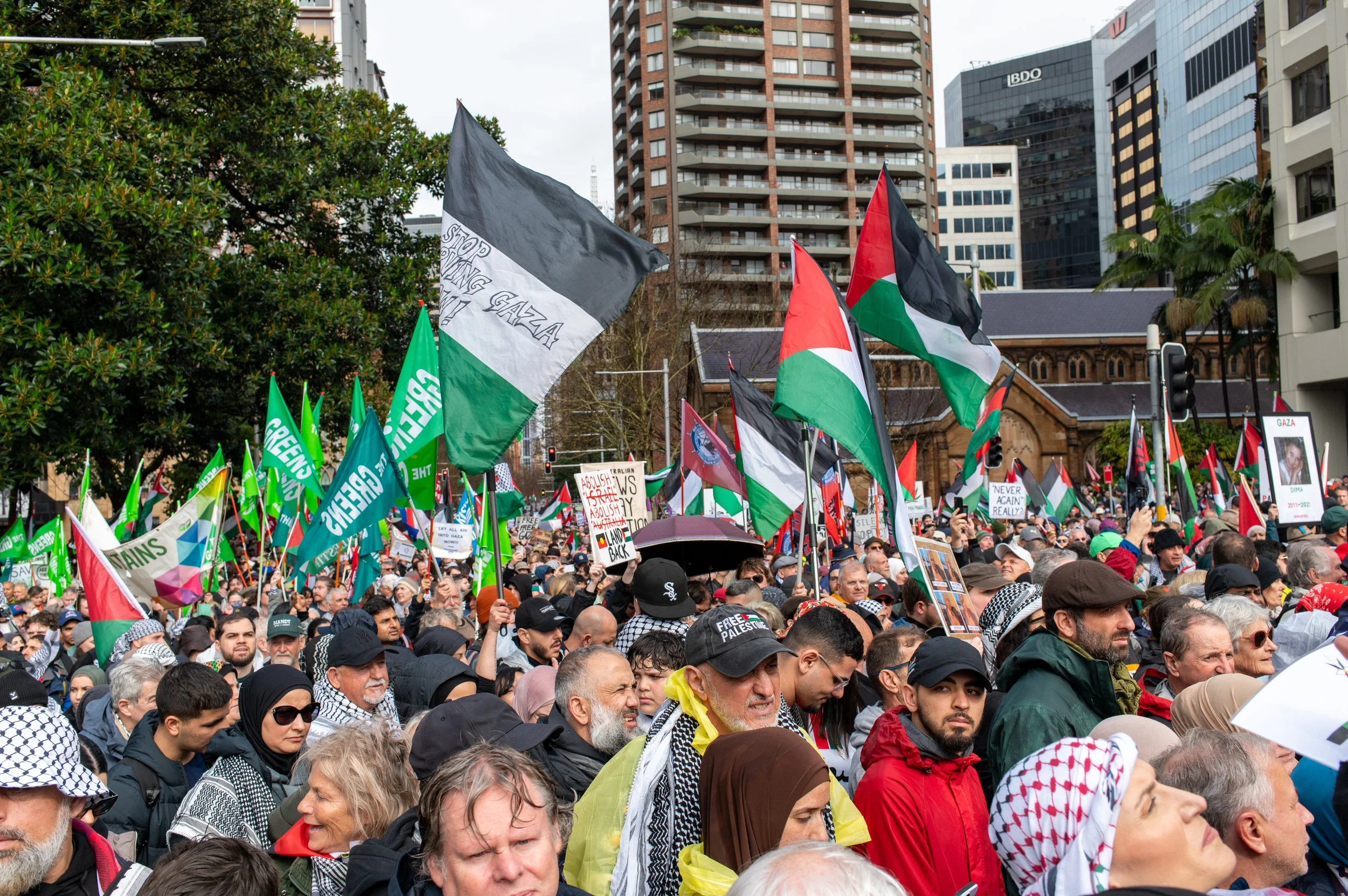 A large crowd participating in a protest or rally, holding various flags including Palestinian flags and green flags supporting Palestine, with some signs in the background. The event is happening outdoors in an urban area with tall buildings and trees.