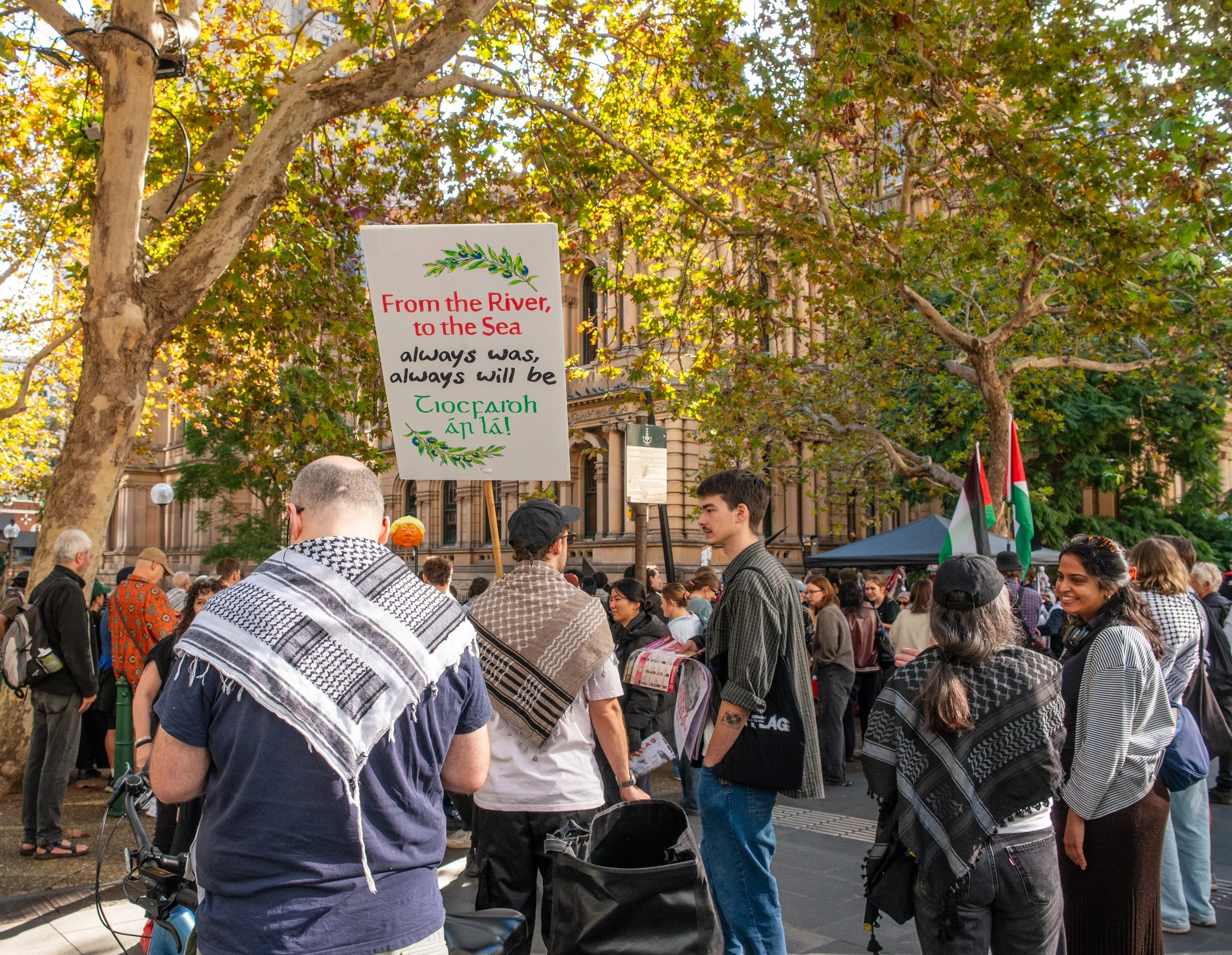 People gathering in a park with trees and historic buildings, holding signs and flags, participating in a demonstration or rally.