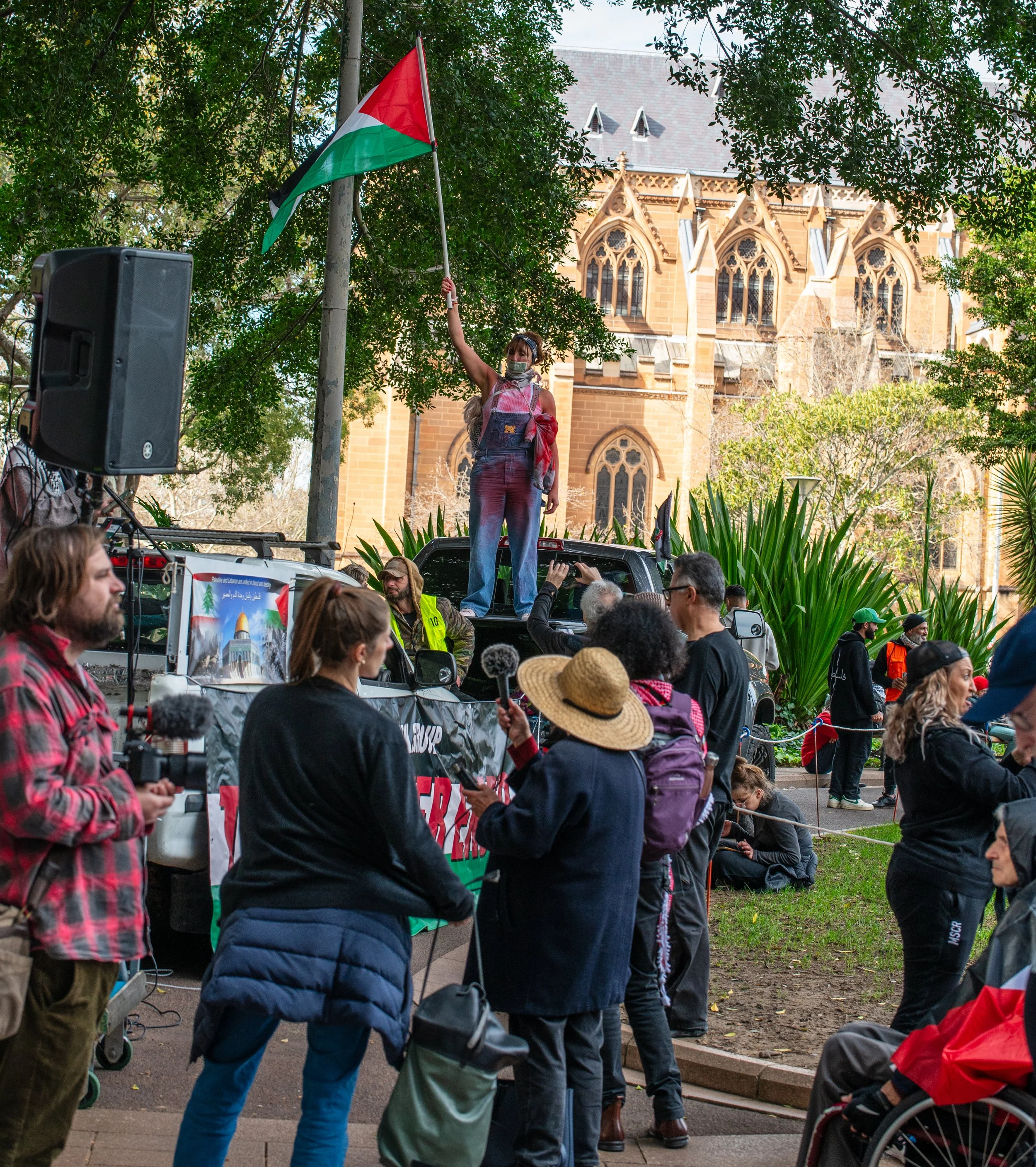 A woman standing on a vehicle holding a Palestinian flag at a protest, surrounded by a crowd of diverse people, some holding smartphones and cameras, with a historic building and trees in the background.