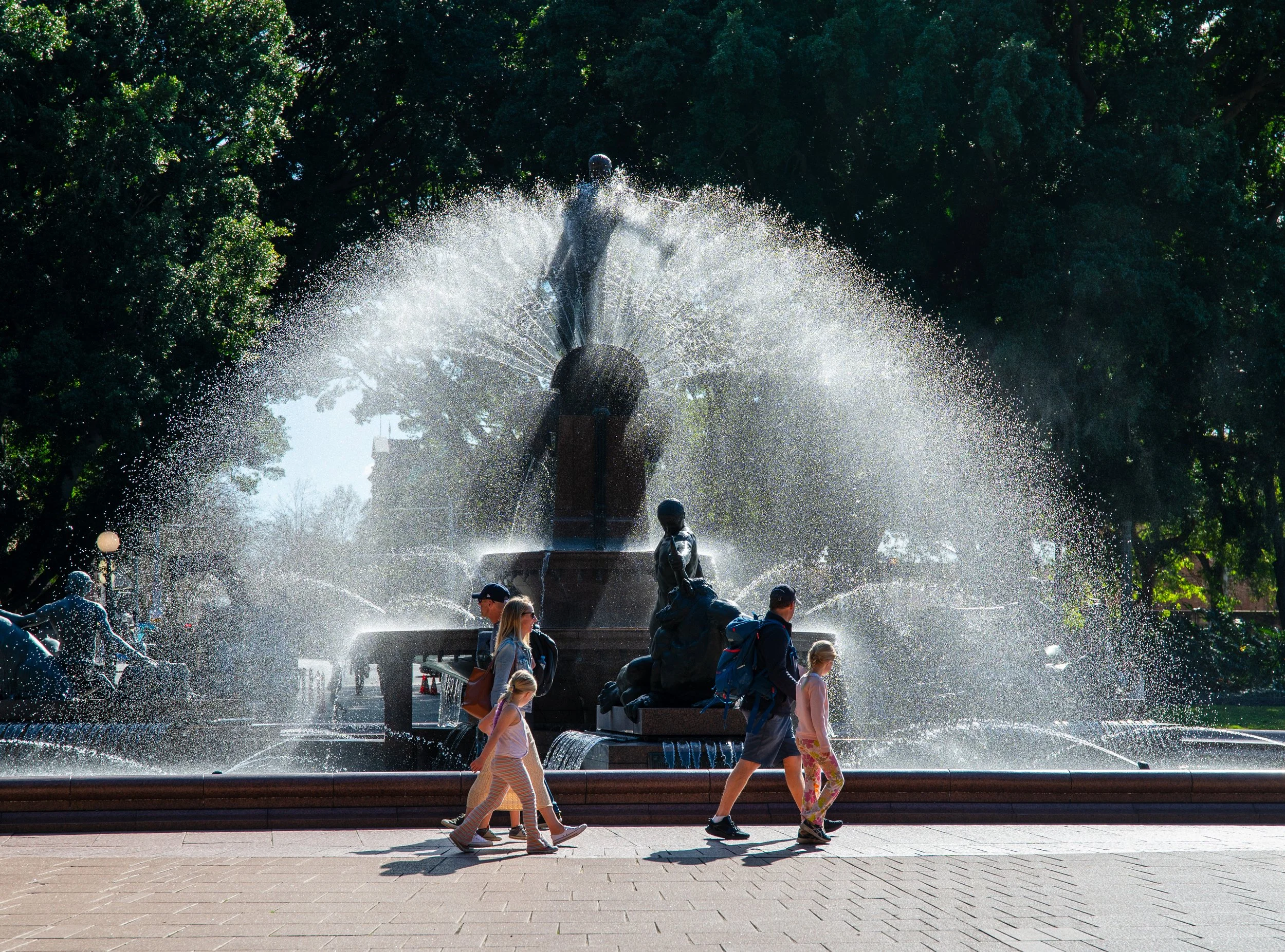 People walking past a large fountain with sculptures and water sprays, surrounded by trees.