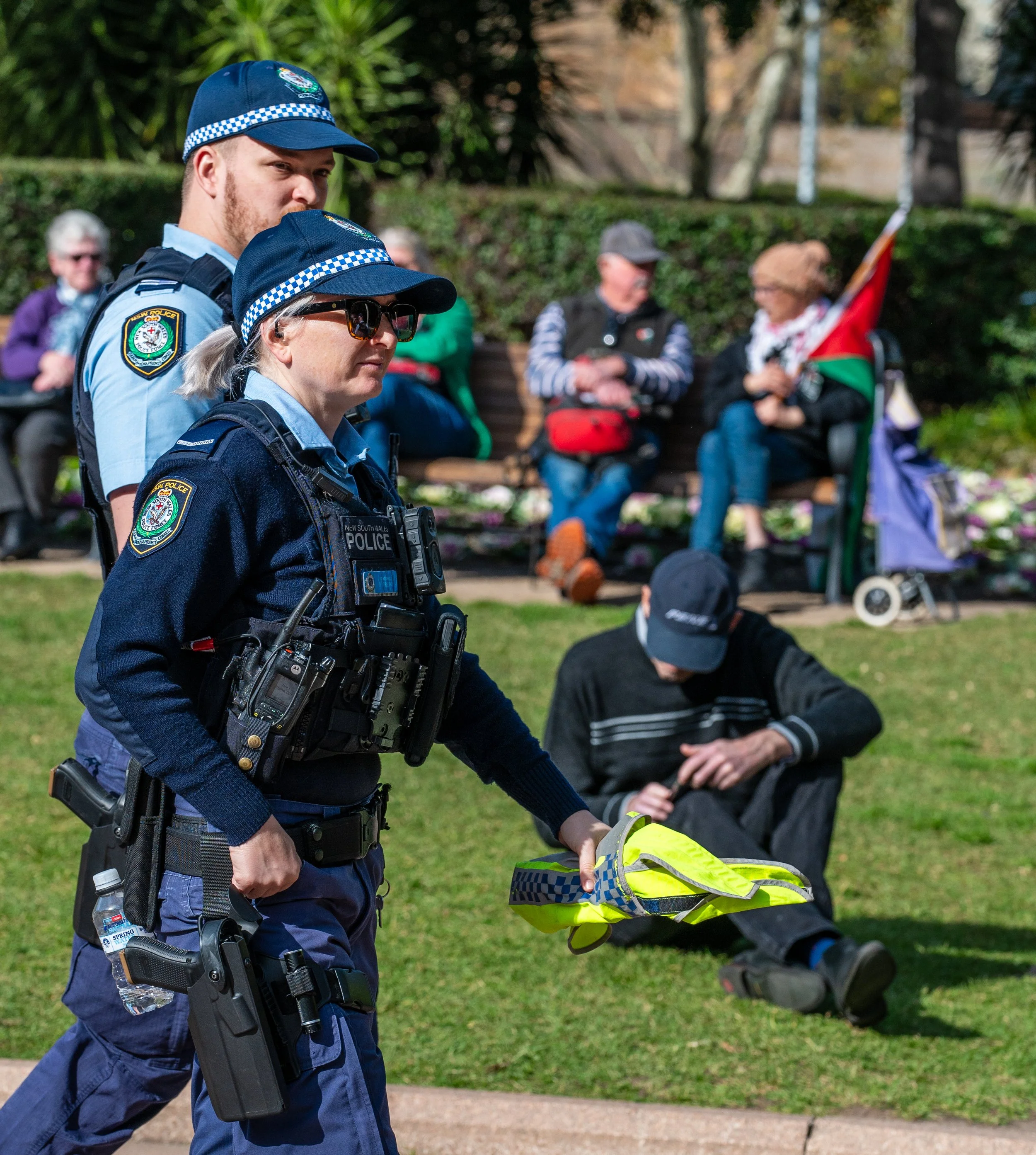 Two police officers, a man and a woman, walking through a park, with people seated on benches and a person sitting on the grass in the background.