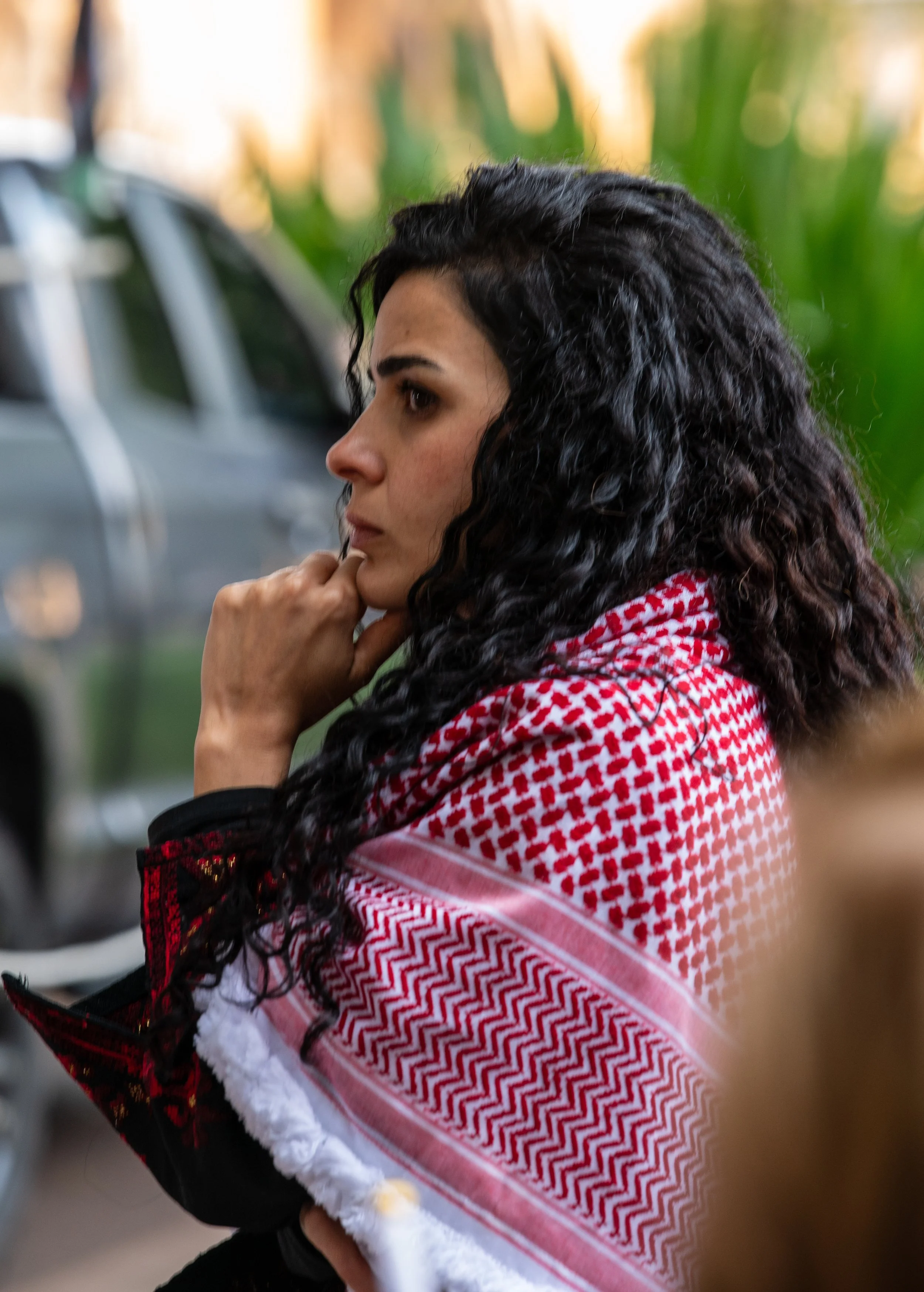 A woman with long curly black hair, wearing a red and white keffiyeh scarf, is resting her chin on her hand and looking thoughtfully to the side outdoors.