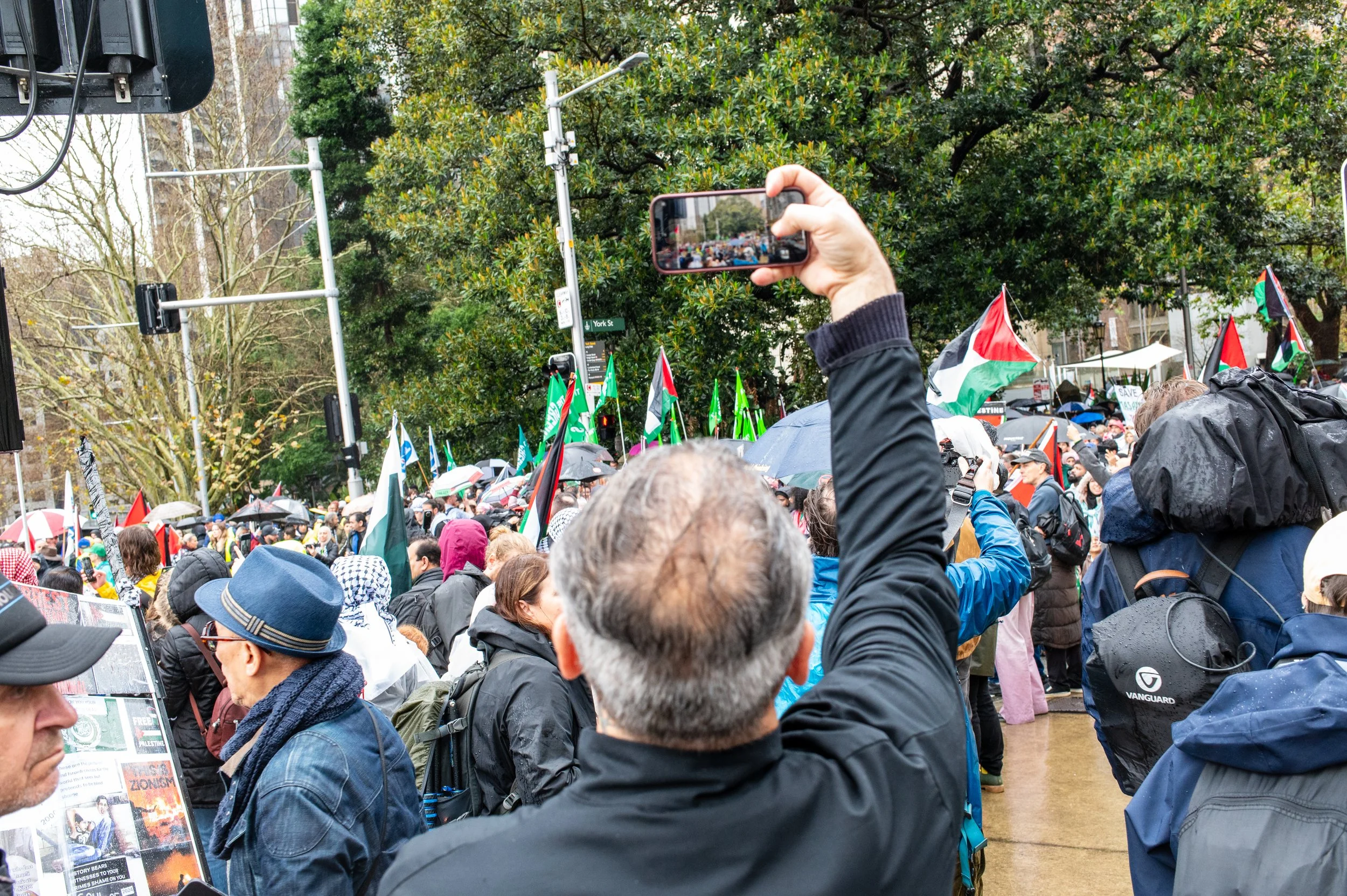 Crowd of people at a protest or rally, many holding umbrellas and flags. A man in the foreground is taking a photo with his phone.