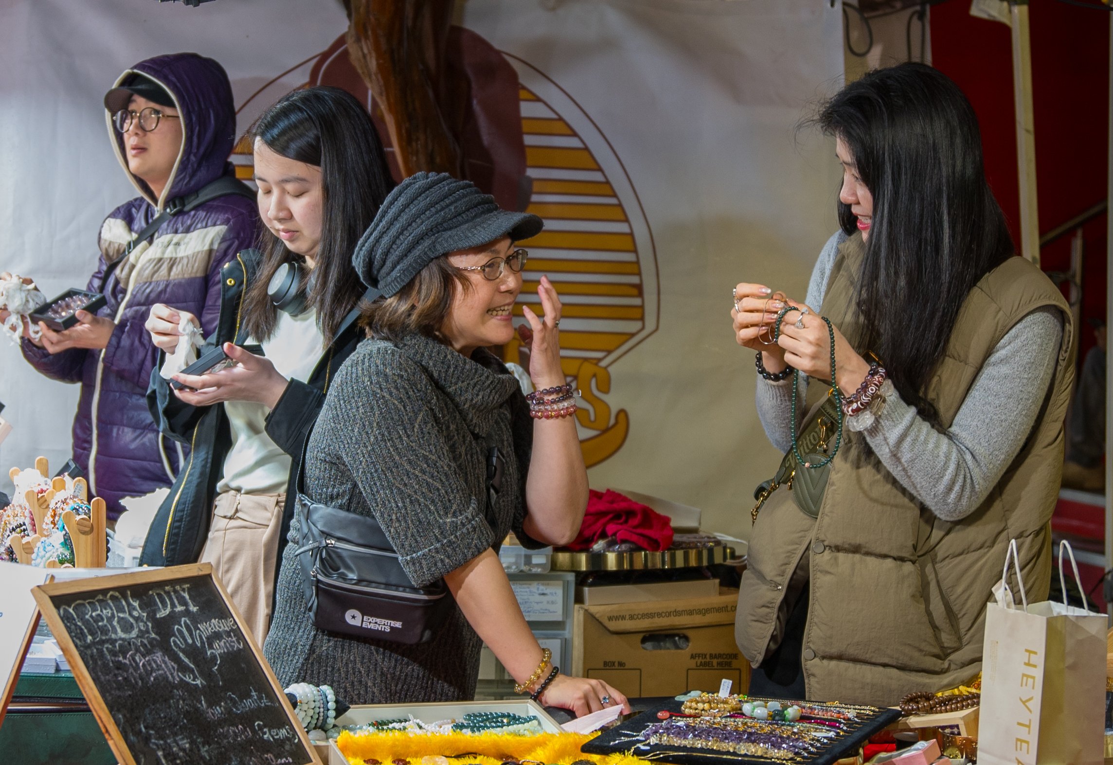 Two women engaging in a lively conversation at a craft stall, surrounded by jewelry and accessories, with two other women looking at items in the background.
