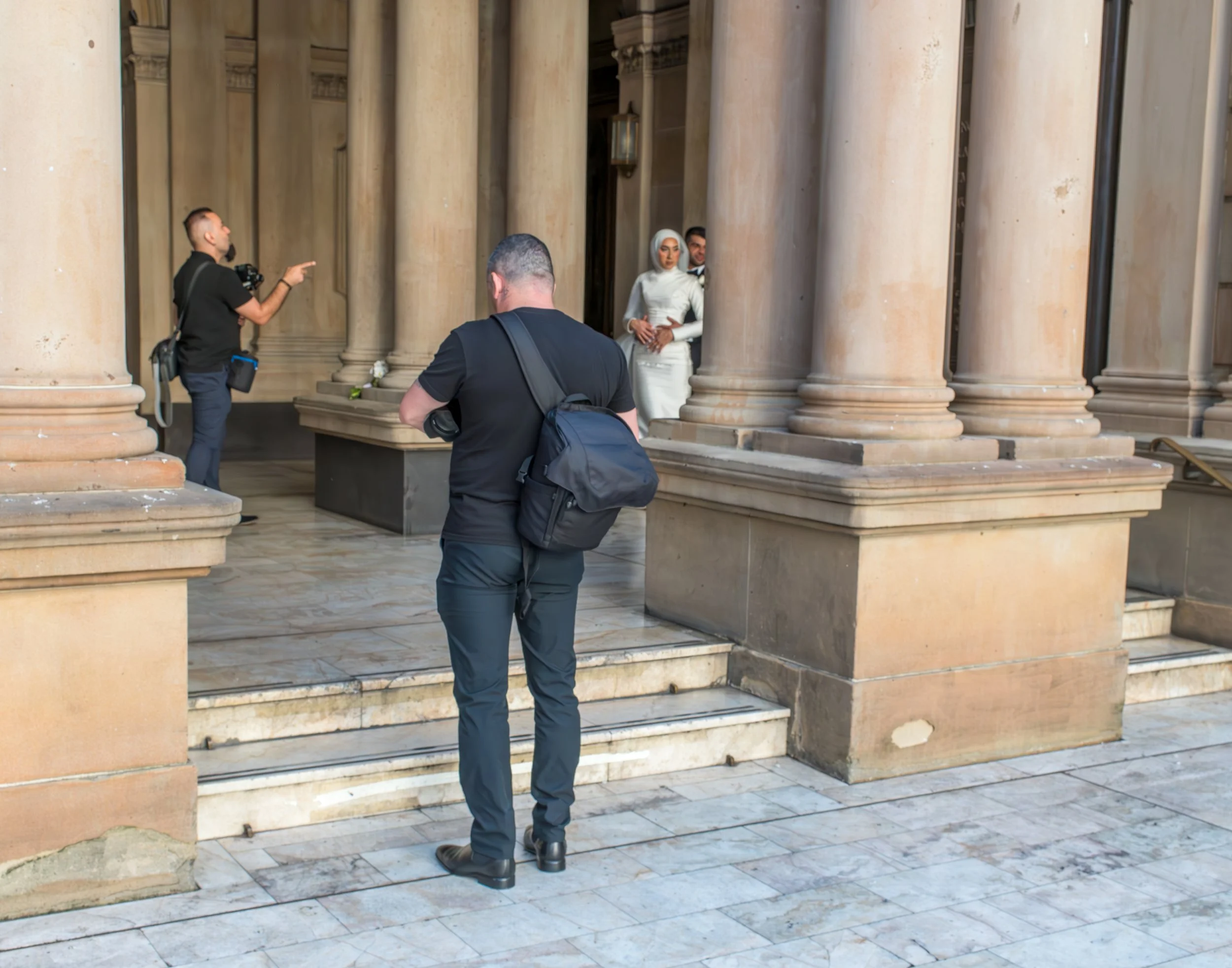 A man with a black shirt and carrying a backpack is standing on steps in front of a grand building with large beige columns, while two other men, one pointing, are engaging with a woman in a white dress and headscarf near the columns.