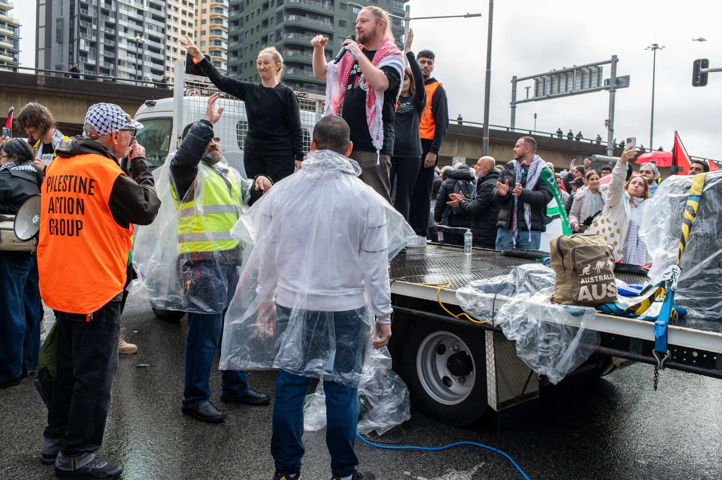 People participating in a protest rally outdoors in an urban setting on a rainy day. Some are standing on a raised platform, speaking or gesturing, while others are listening. Many attendees are wearing rain ponchos, and some are holding flags and banners.