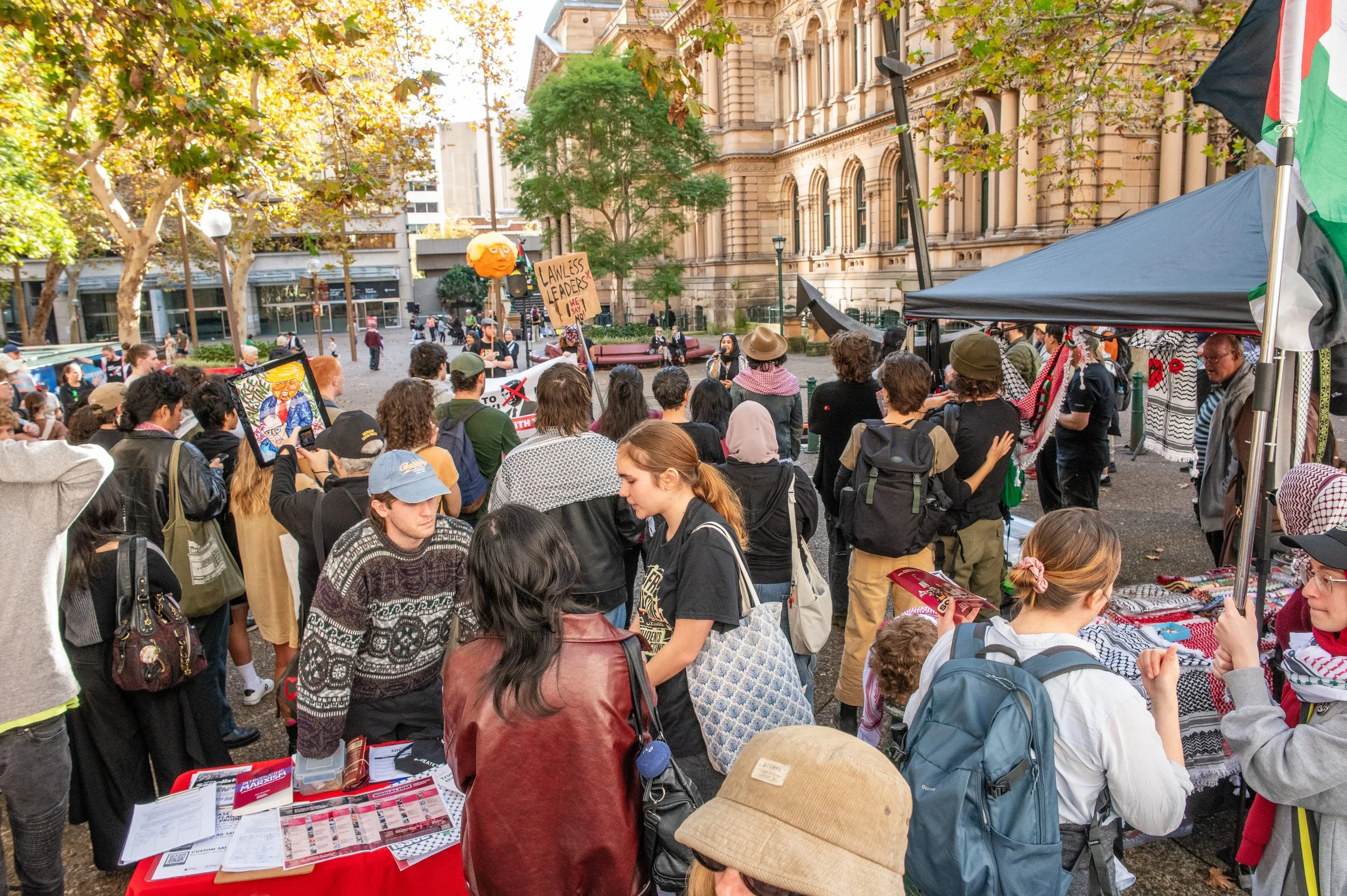 Crowd gathered at a protest or rally in an urban park with buildings in the background, some holding signs and flags, others browsing stalls, and a puppet resembling a caricature of a person, with trees and sunlight highlighting the scene.