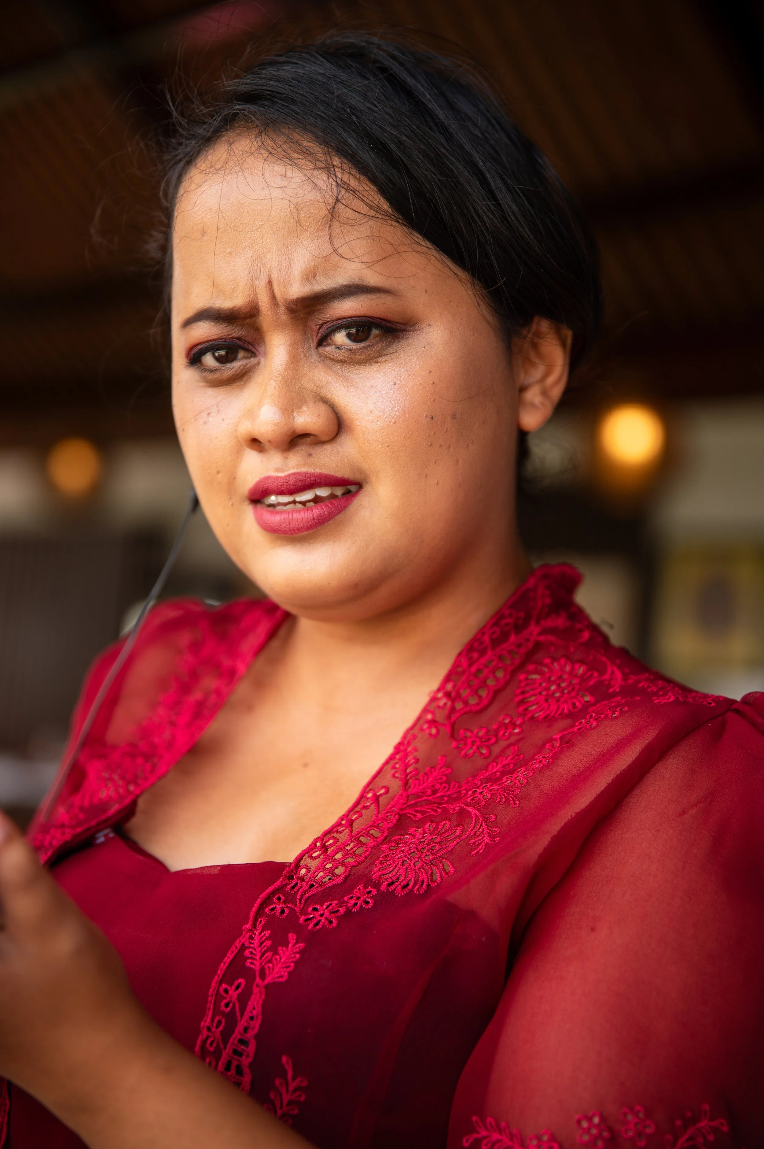 A woman in a red embroidered dress looking at the camera, with a serious expression, against a blurred, warm background.