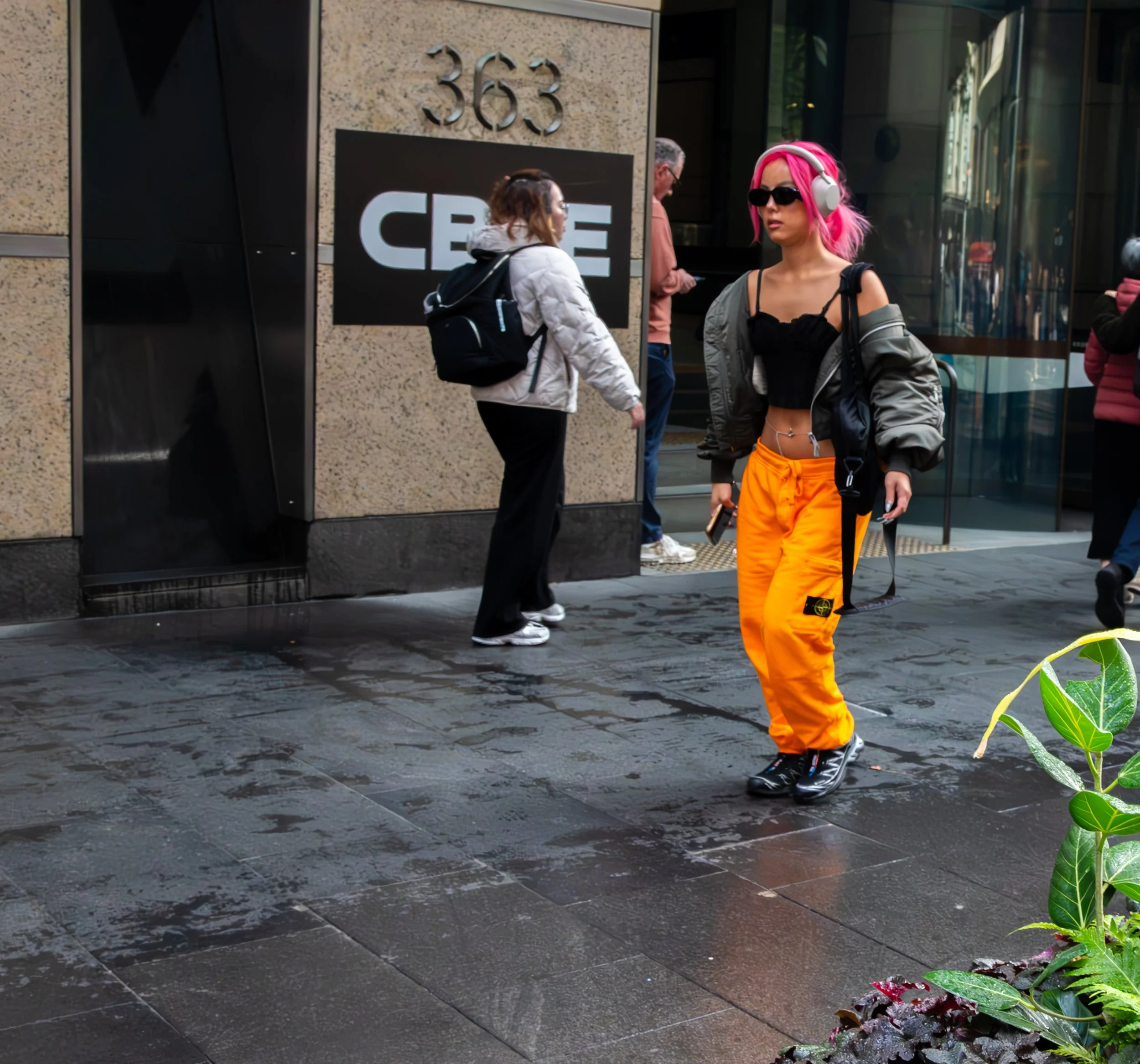 A woman with pink hair, sunglasses, headphones, black crop top, gray jacket, and orange pants walking on a city street.