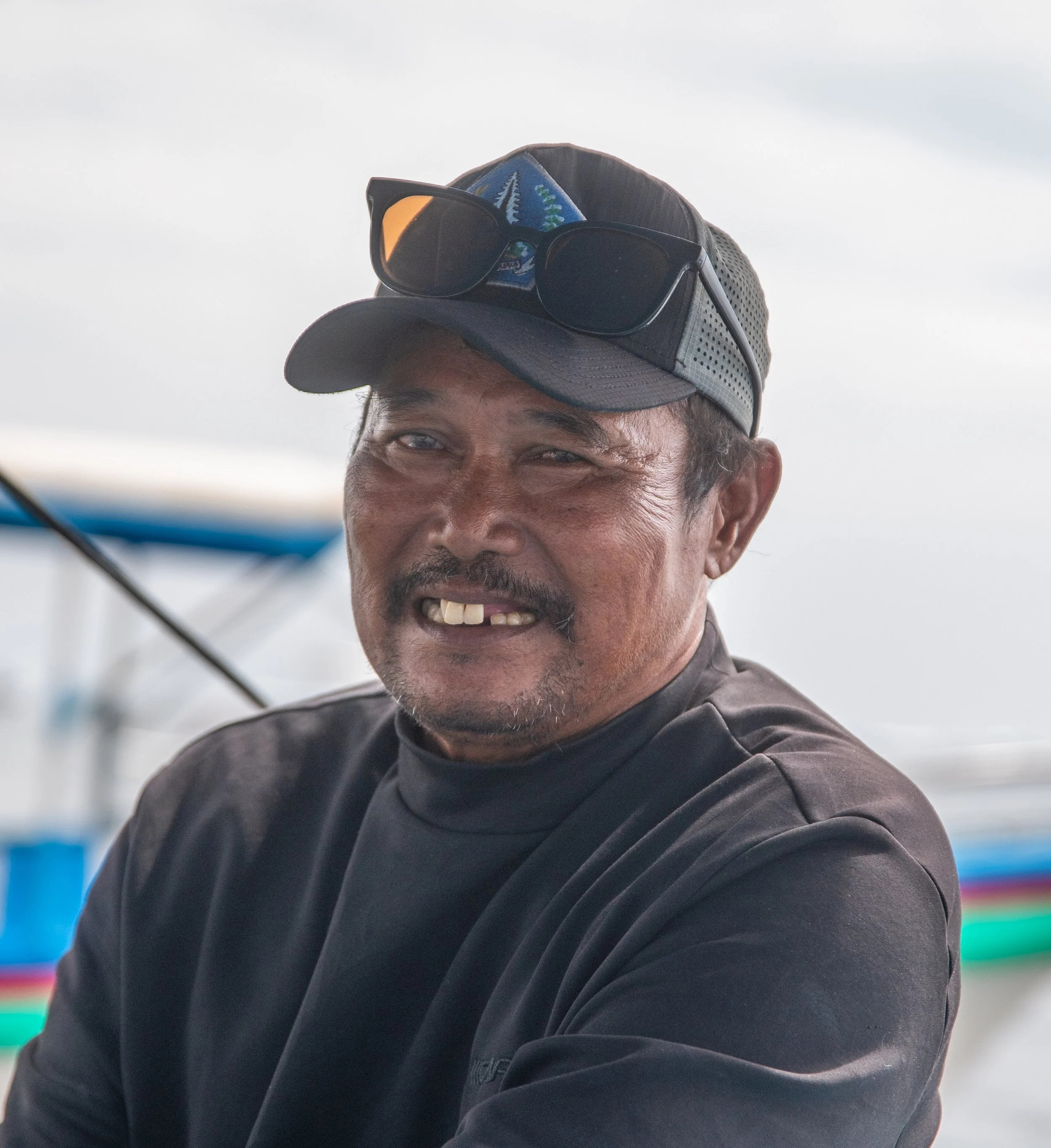 Close-up of a smiling man wearing a cap and sunglasses, with a colorful, blurred background.