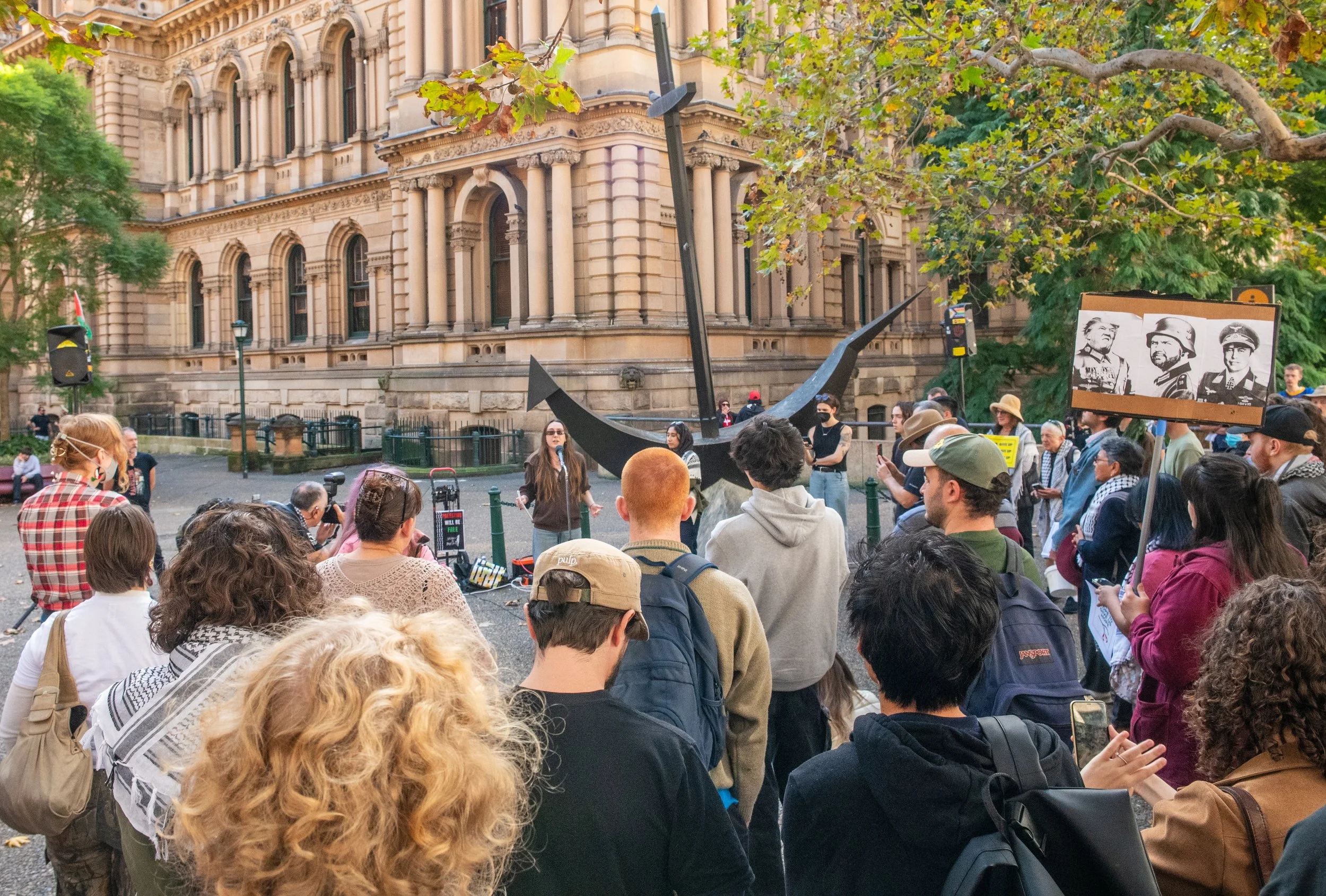 A diverse group of people gathered outdoors in front of a large historic building with beige stone and arched windows. A woman stands in the center speaking to the crowd, with a large anchor sculpture behind her. Some people hold signs, including one with black and white photos of soldiers. The scene is set in autumn with green and yellow leaves on nearby trees.