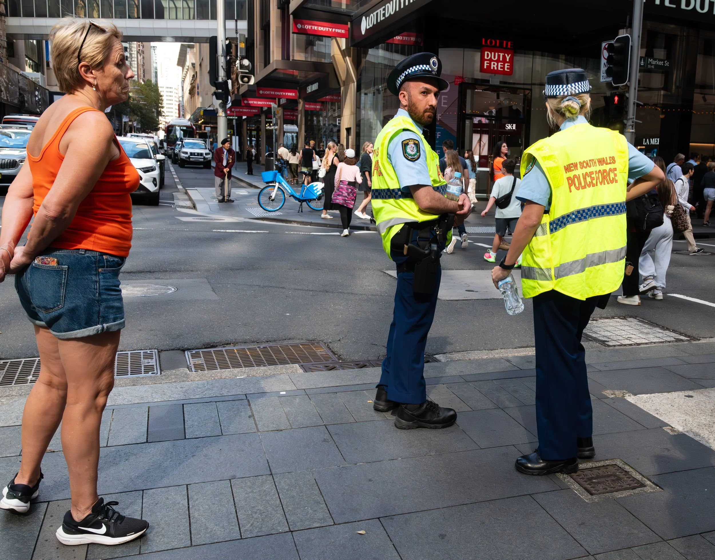 A woman in a red tank top and denim shorts stands on a city sidewalk, facing two police officers from the New South Wales Police Force, who are wearing yellow high-visibility vests. The officers are engaged in conversation, with the female officer holding a water bottle. Pedestrians and vehicles are visible in the background at a busy city intersection.