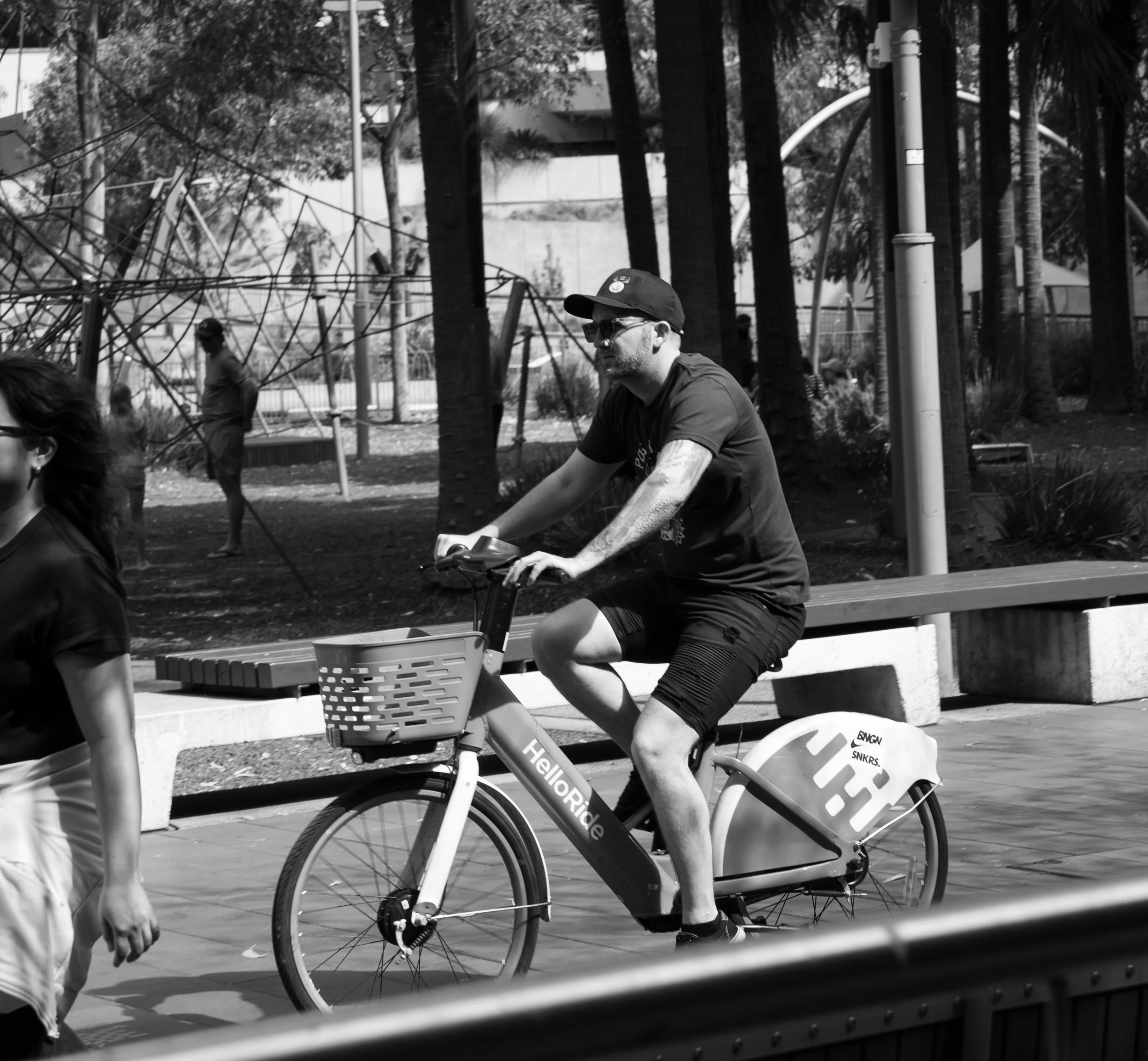 A man riding an electric bike on a sidewalk in a park with trees, a playground, and benches in the background during daytime.