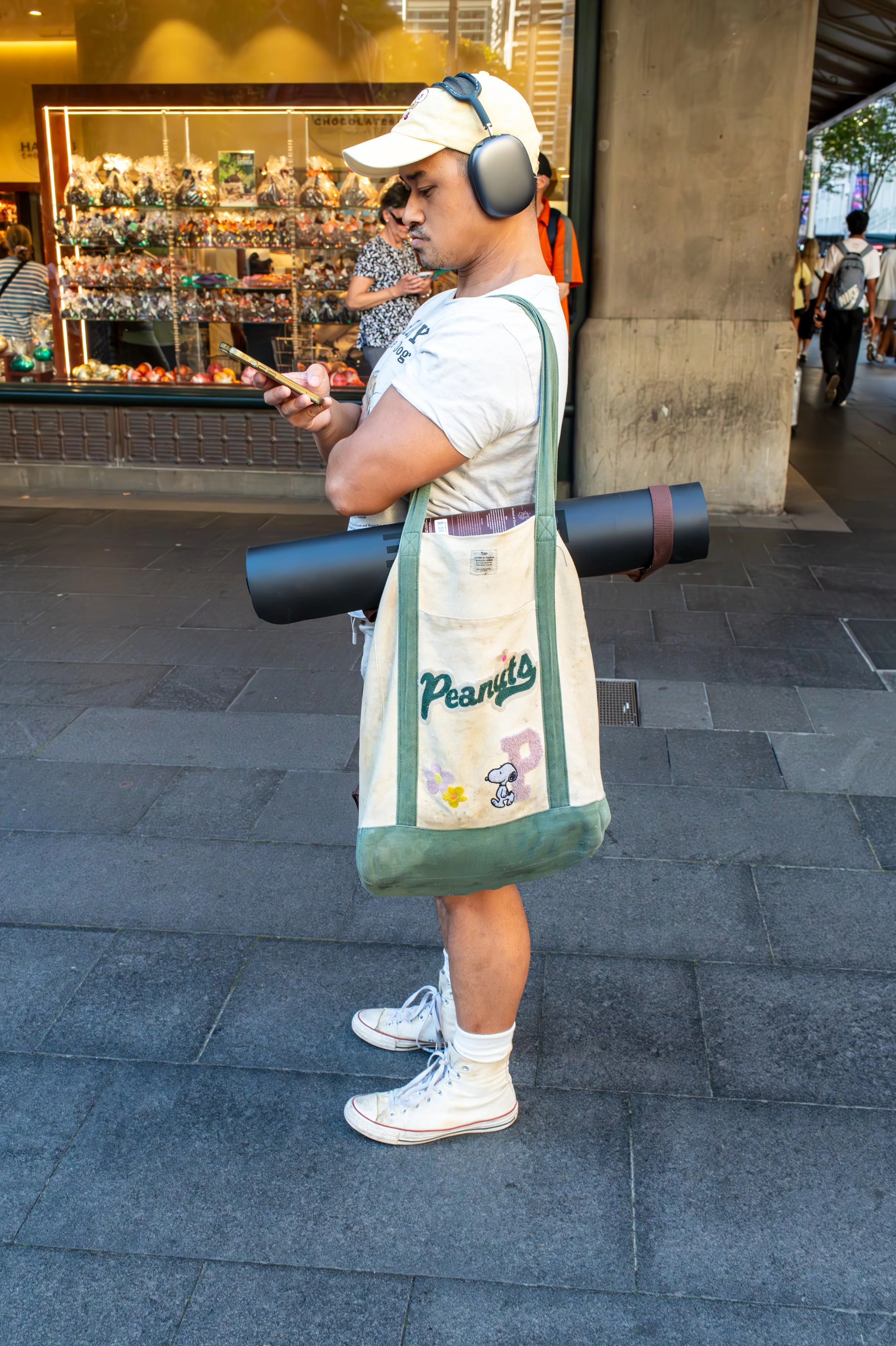 Young man with headphones, beige cap, white t-shirt, white high-top sneakers, carrying a Peanuts Tote Bag, and a rolled-up yoga mat, standing on a city sidewalk and looking at his phone.