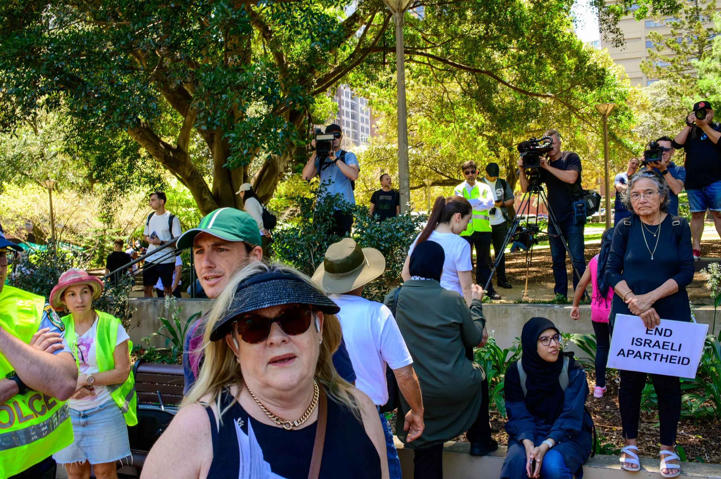 A woman holding a sign that reads 'End Israeli Apartheid' at a protest in a park with people, cameras, and police officers around.