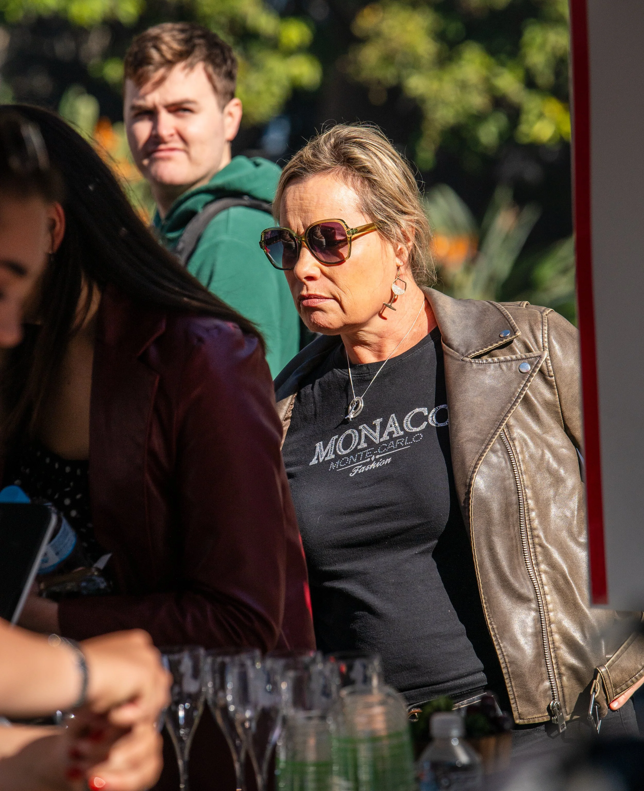 A woman wearing sunglasses, a black t-shirt with 'Monaco Monte Carlo' printed on it, and a brown leather jacket looking at a display or counter. A young man in a green hoodie stands behind her, and part of another person in a maroon jacket is also visible in the foreground.