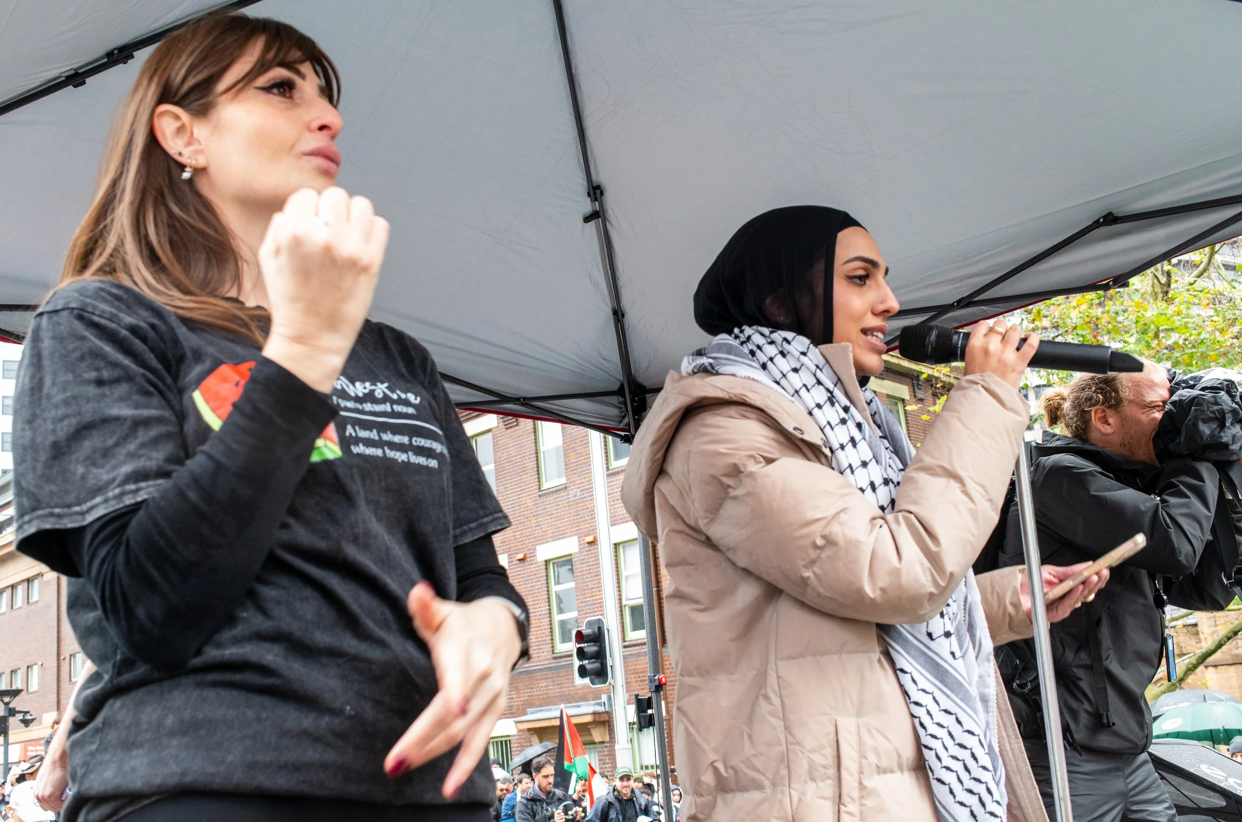 Two women speaking and listening at a crowd event under a tent, one holding a microphone and the other with a cellphone, with people and buildings in the background.