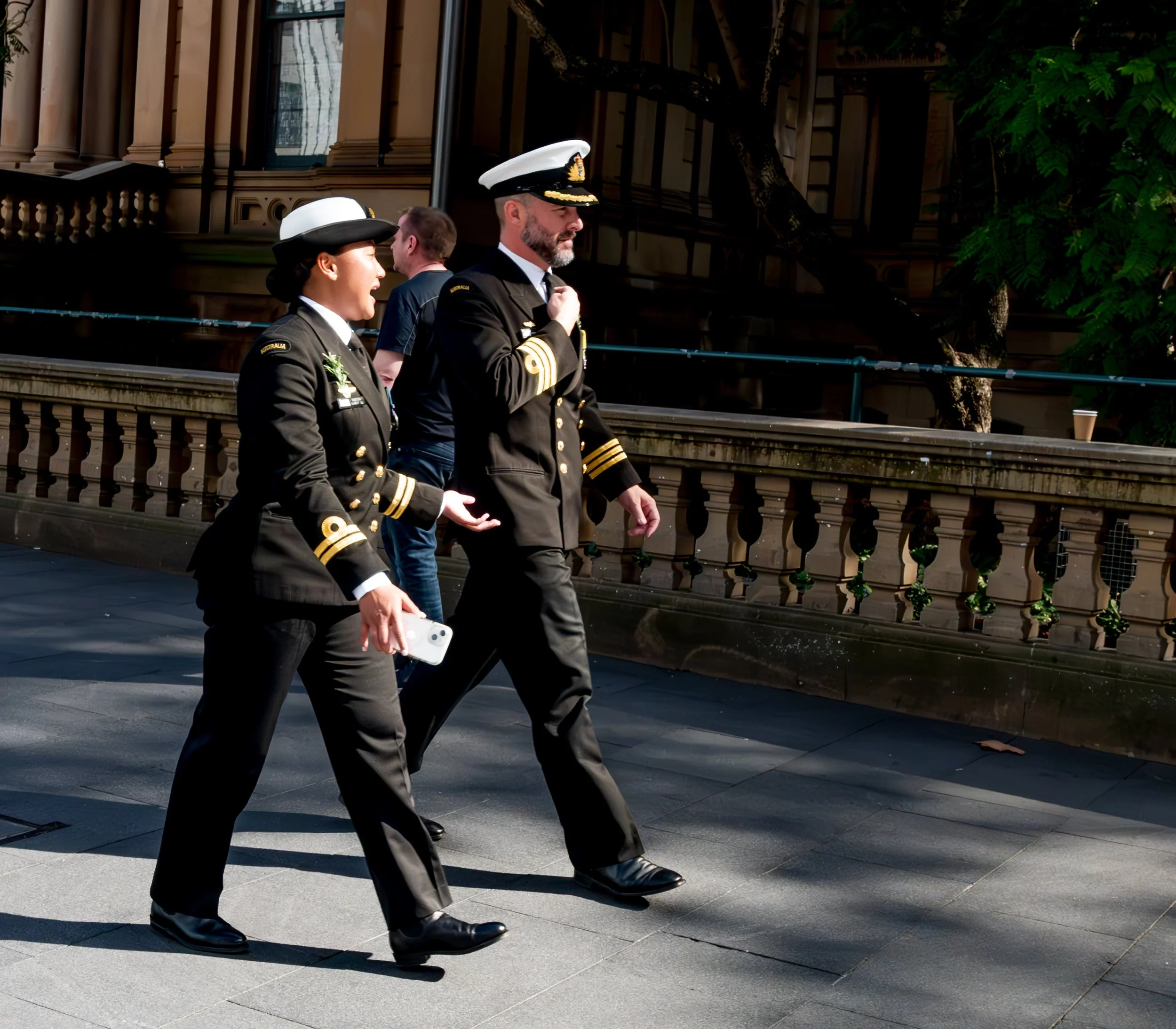Two naval officers, a woman and a man, walking together on a city sidewalk, dressed in formal navy uniforms with hats and gold insignia, with a building and trees in the background.