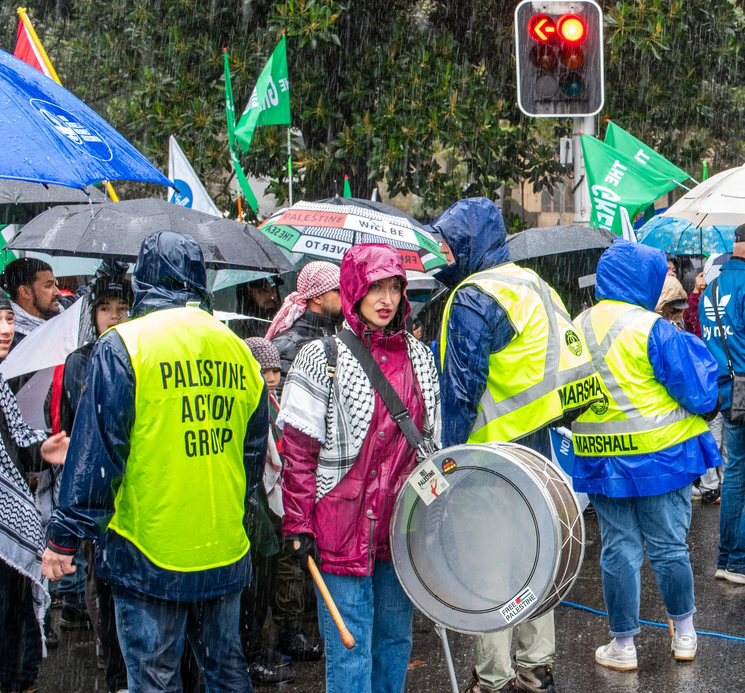 Crowd of protesters in the rain, some with umbrellas, participating in a demonstration holding flags and banners, with a woman in front, wearing a pink raincoat and Arabic scarf, surrounded by others wearing yellow vests. Traffic lights are visible in the background.