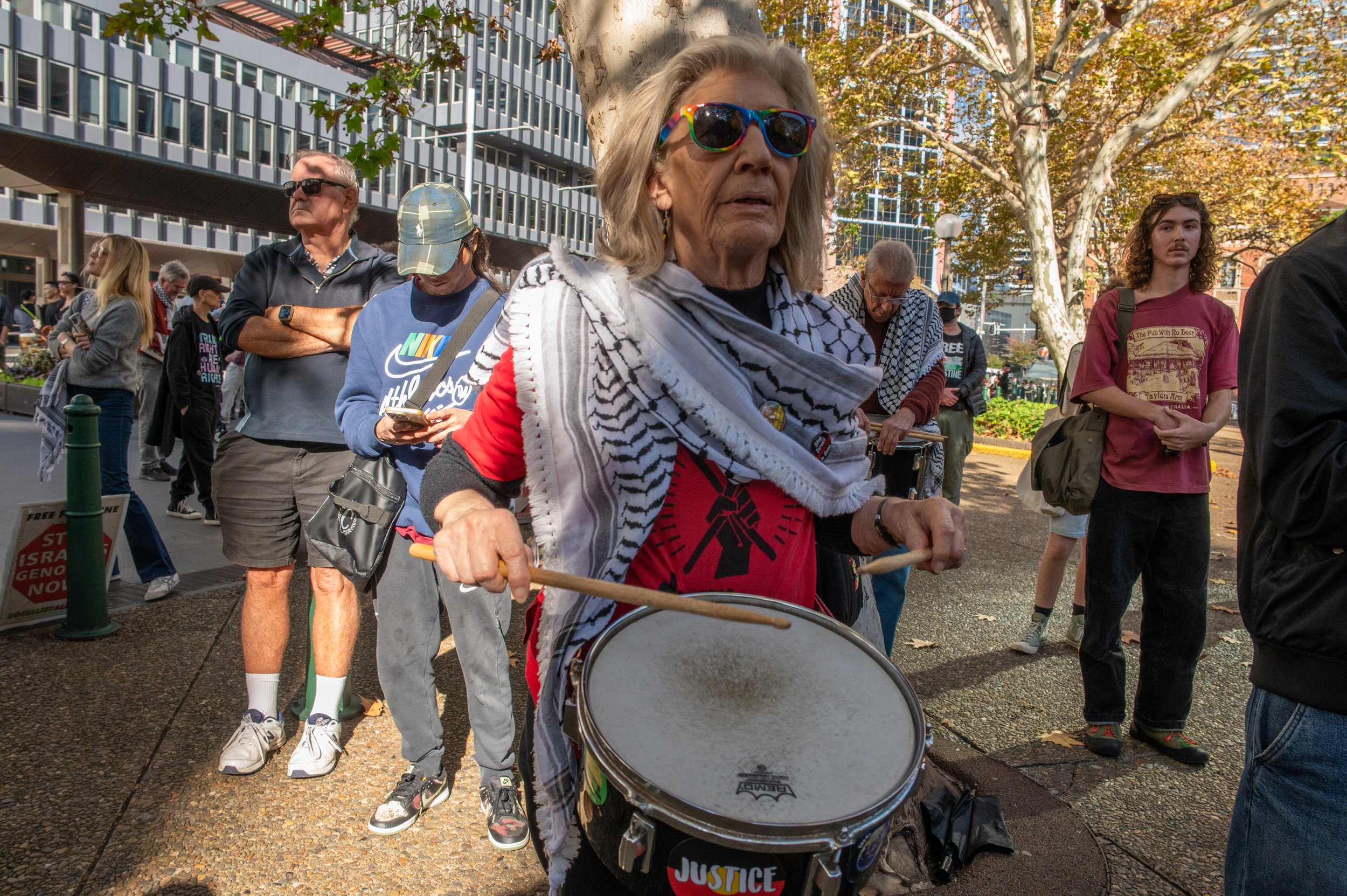 An elderly woman wearing sunglasses and a keffiyeh scarf playing a drum at a protest or rally, surrounded by diverse people standing in a city park with trees, buildings, and a sidewalk.