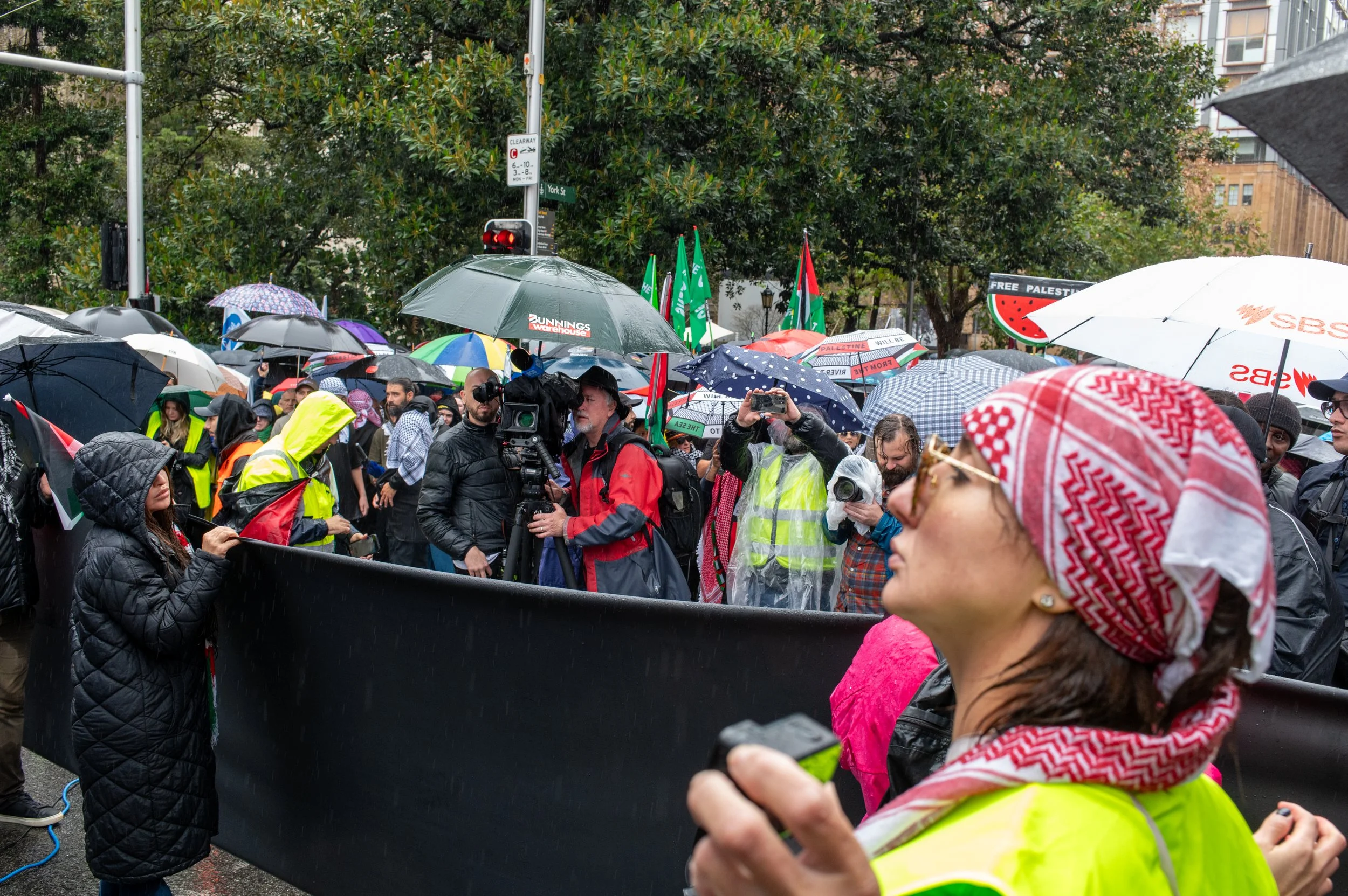 A crowd gathering at a protest in the rain, with many people holding umbrellas and signs, some wearing yellow vests and raincoats, amidst media presence and flags.