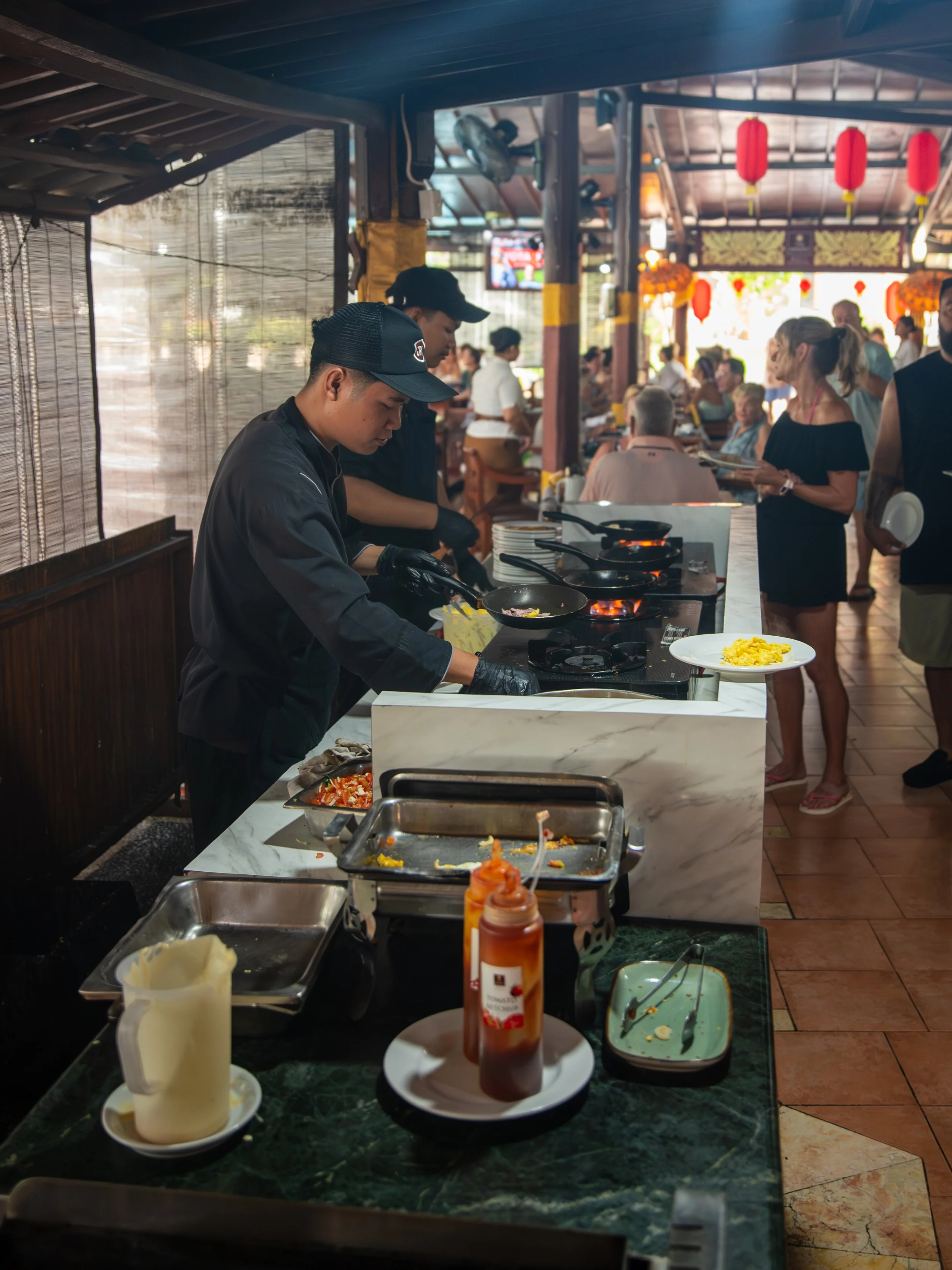 A busy restaurant kitchen with chefs preparing food at a marble-topped counter, surrounded by customers and decorated with red lanterns and yellow flowers.