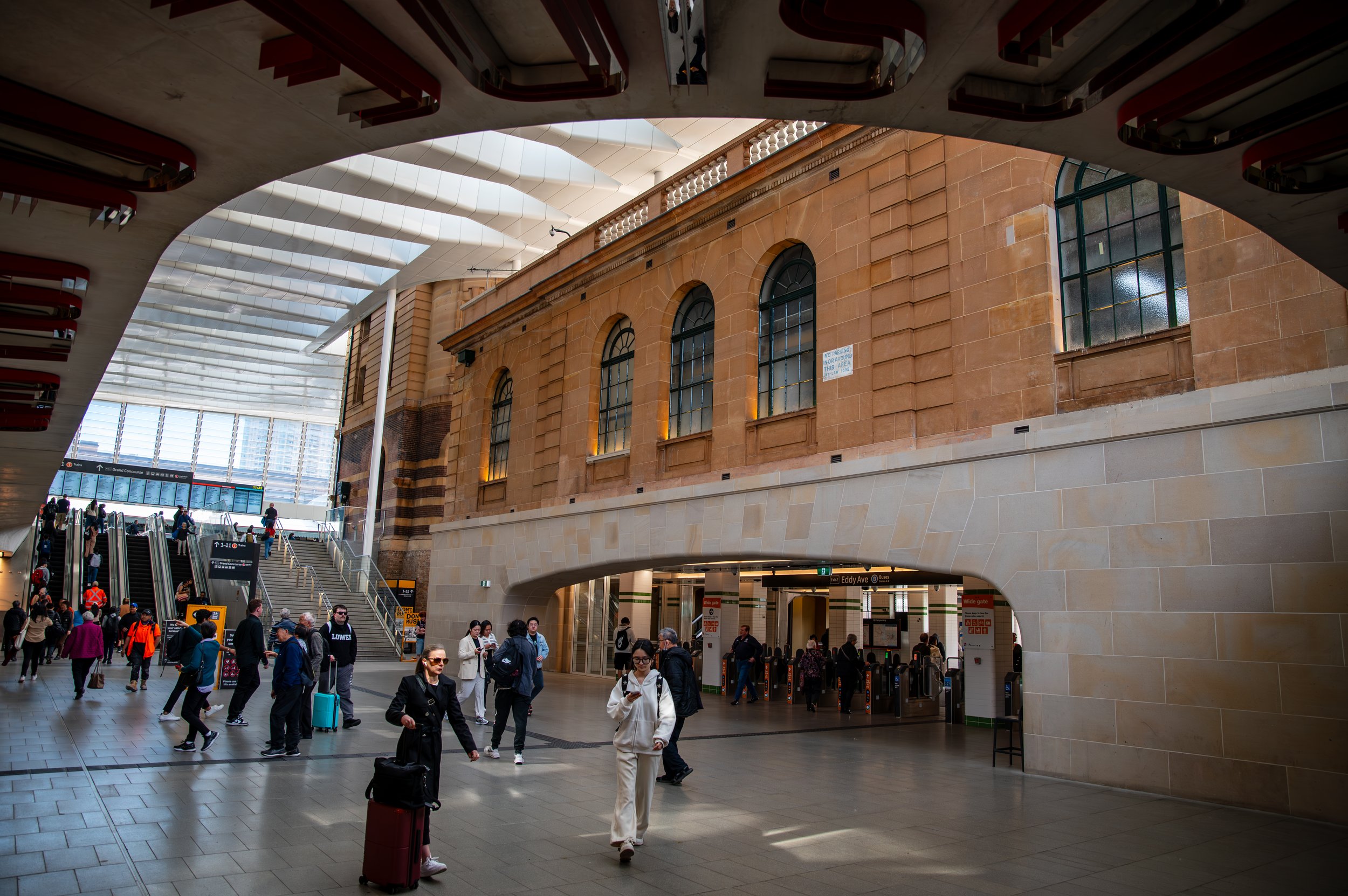 Inside a busy train station with people walking and carrying luggage, featuring escalators, modern architecture, a textured ceiling, and historical brick building elements.