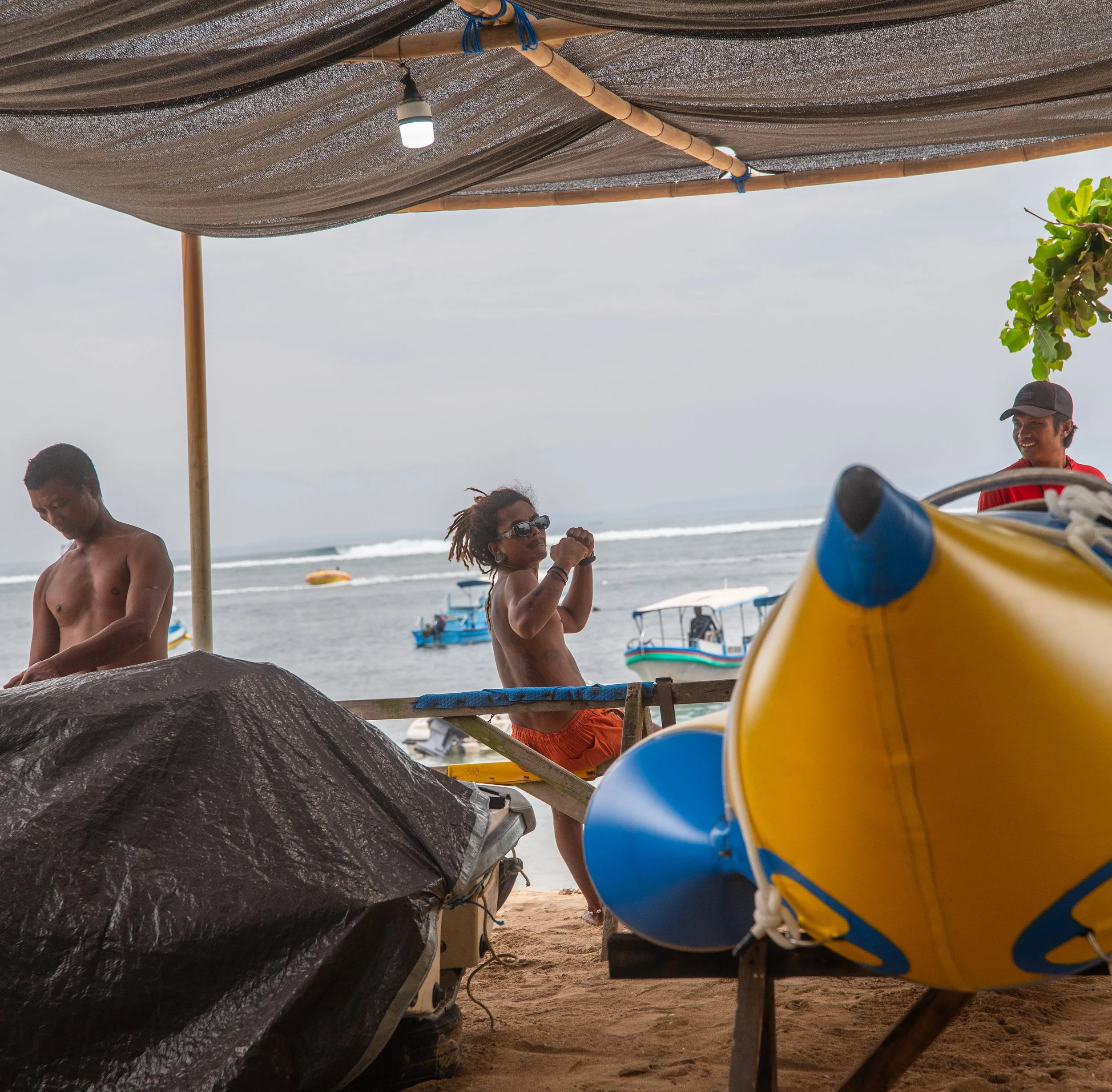 Beach scene with a shade canopy made of fabric and bamboo poles, with people standing and chatting near colorful kayaks on sandy ground, boats floating in the water, and a man wearing a hat smiling.