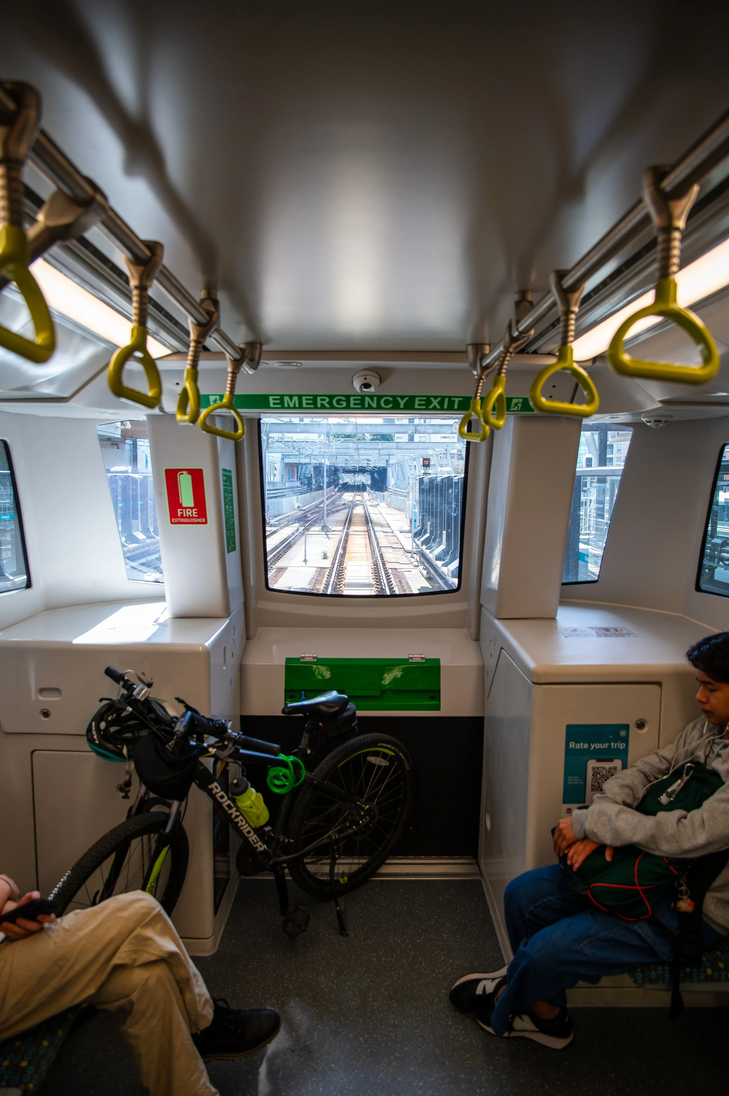 Inside a train with a bicycle parked near the front window, two passengers sitting on seats, hand straps hanging from the ceiling, and view of train tracks ahead.