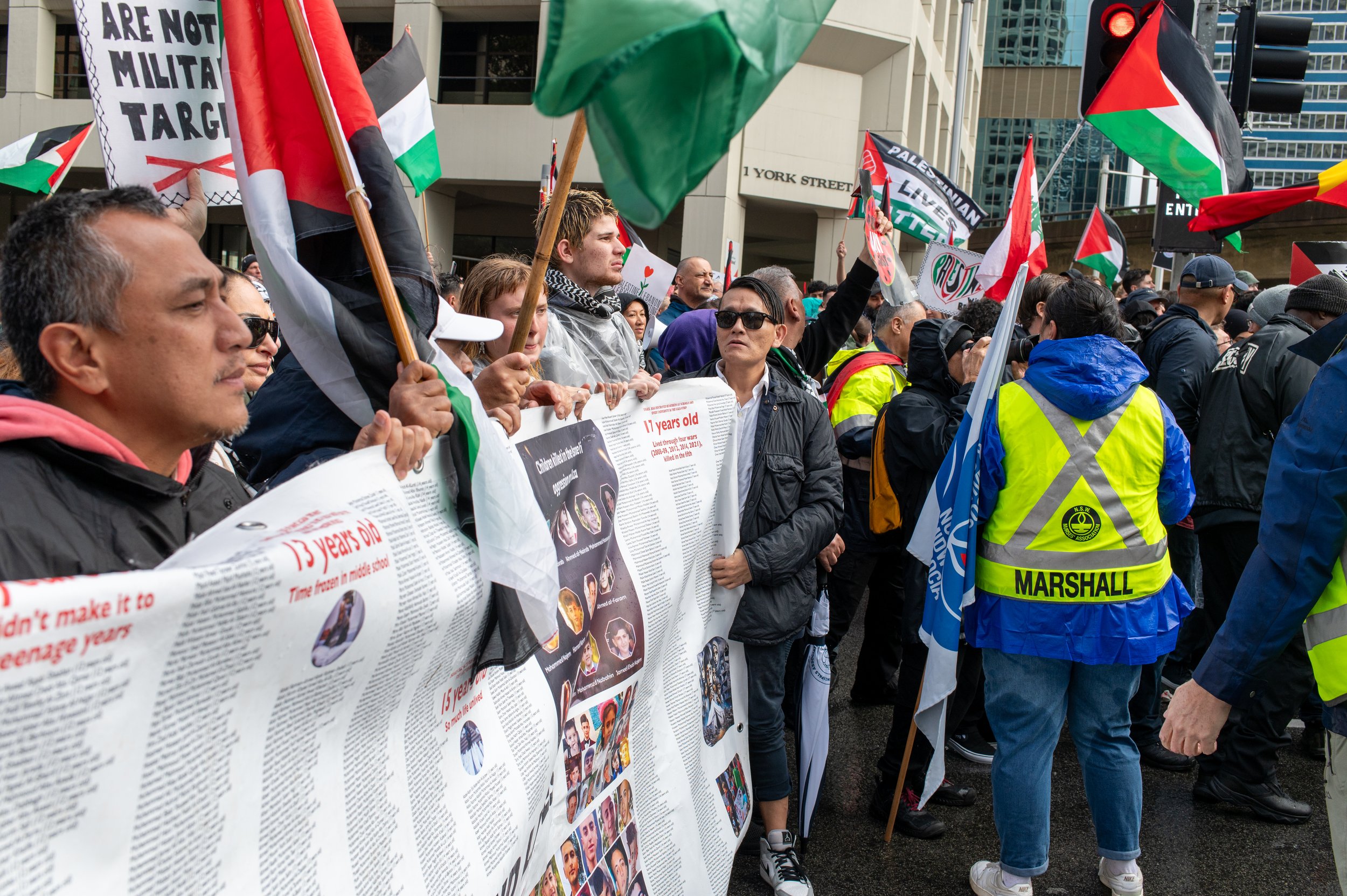 A crowd of protesters holding flags, banners, and signs with images of young victims. Many are wearing jackets, some with a