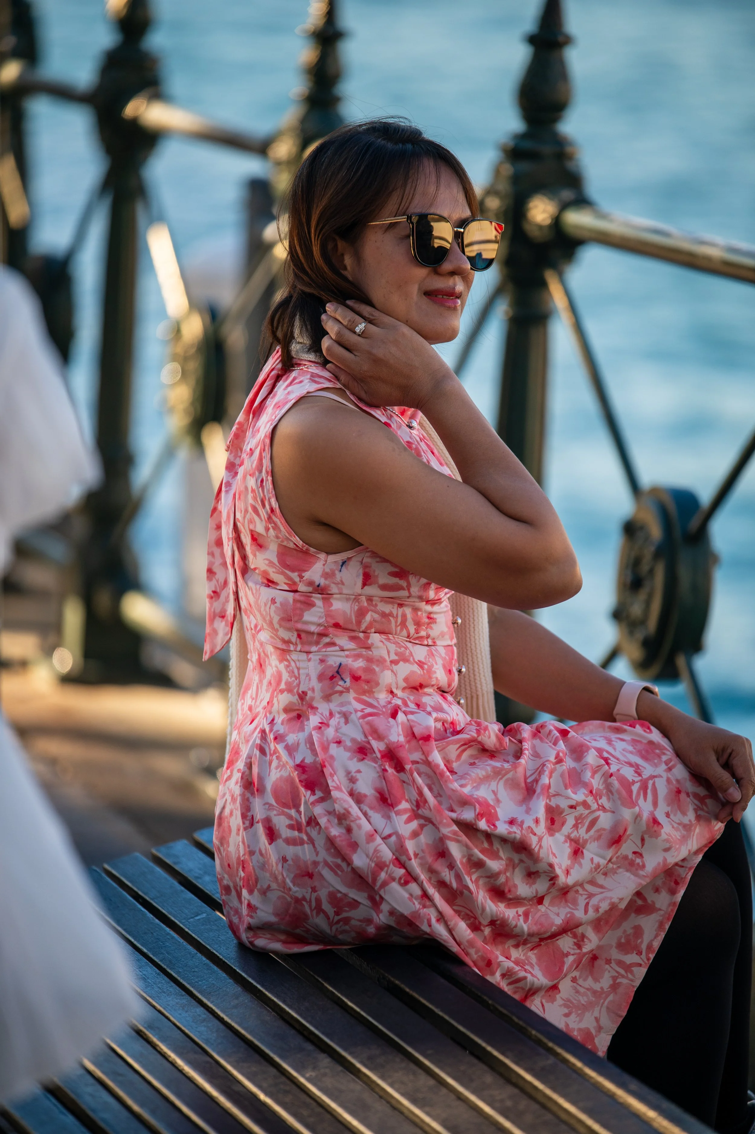 Woman in pink floral dress sitting on a bench by the water, wearing sunglasses and a watch, with a scenic waterfront background.