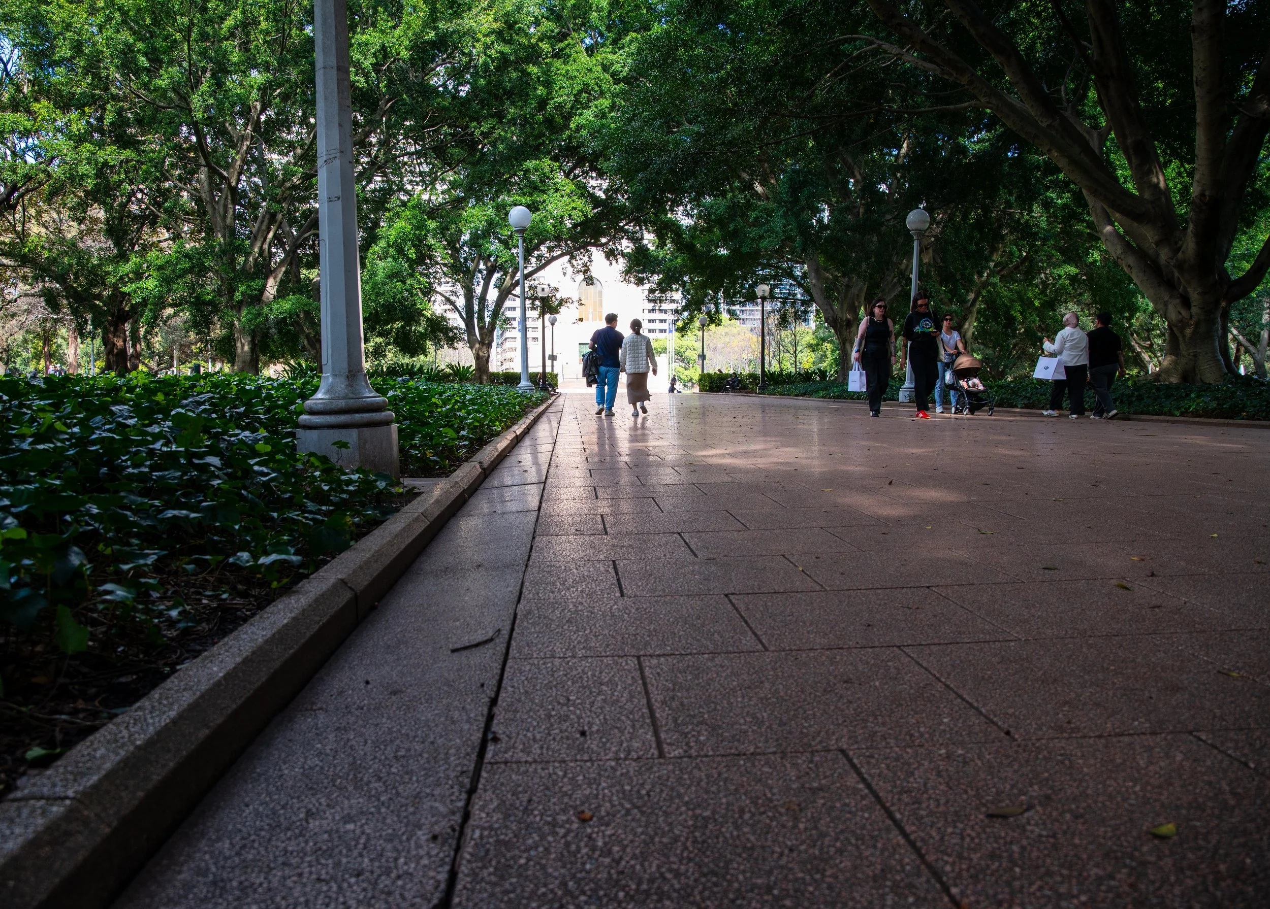 People walking through a shaded park with large trees and lampposts on a paved path.