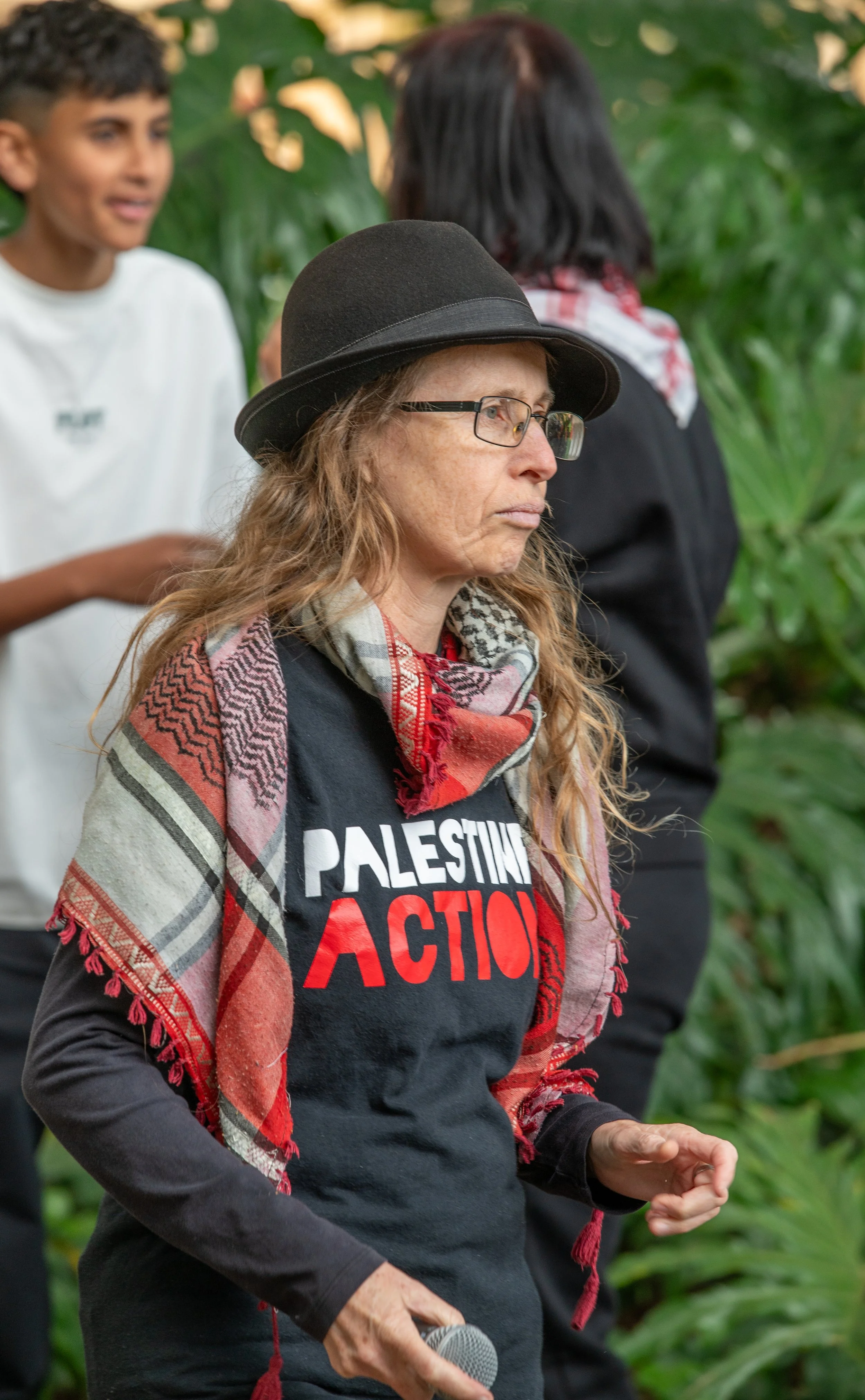 Woman with long hair, glasses, and a black hat holding a microphone, wearing a black shirt with red and white text reading 'Palestine Action', and a patterned scarf, standing outdoors with greenery in the background. Two children are blurred in the background.