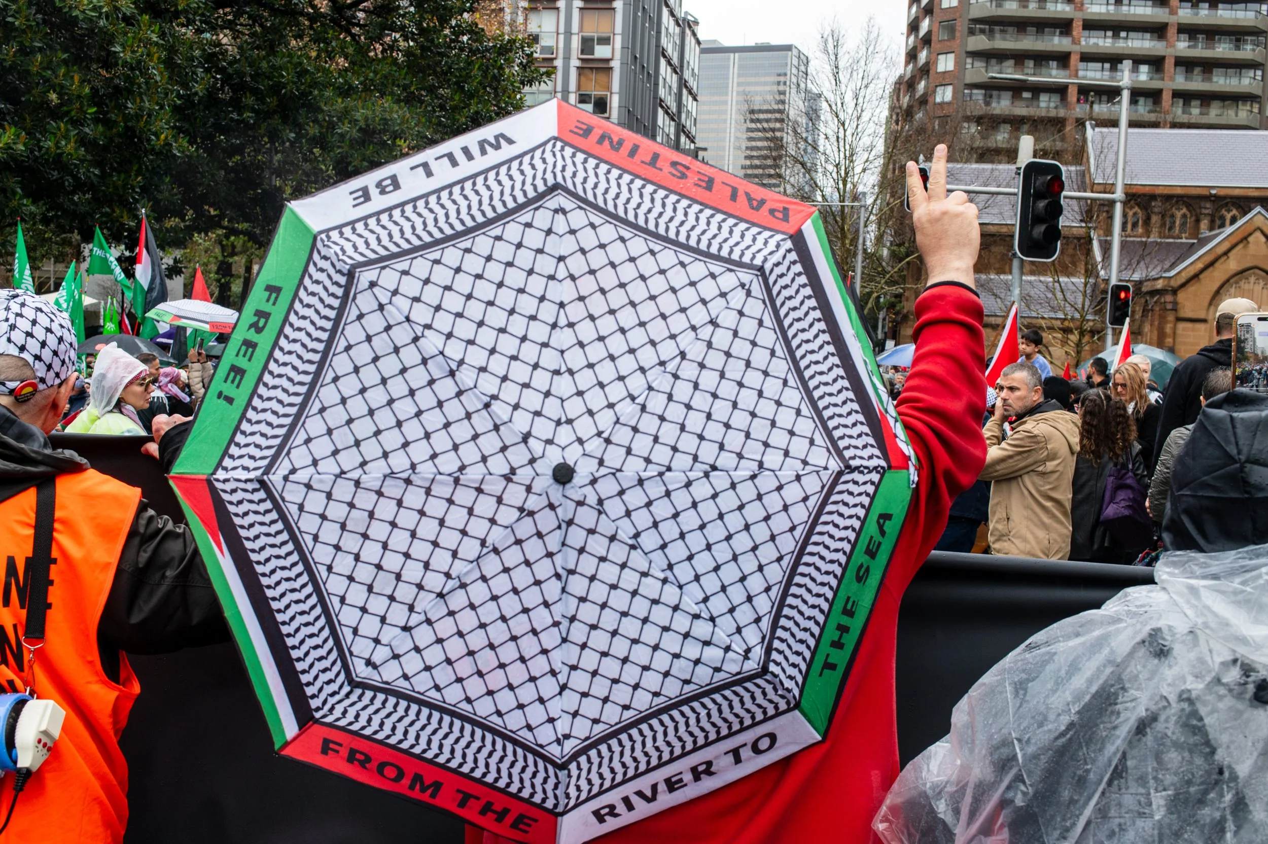 Crowd of people protesting with flags, some wearing rain gear, in an urban area with buildings and traffic lights, holding umbrellas, including a close-up of a umbrella with a geometric pattern and text.