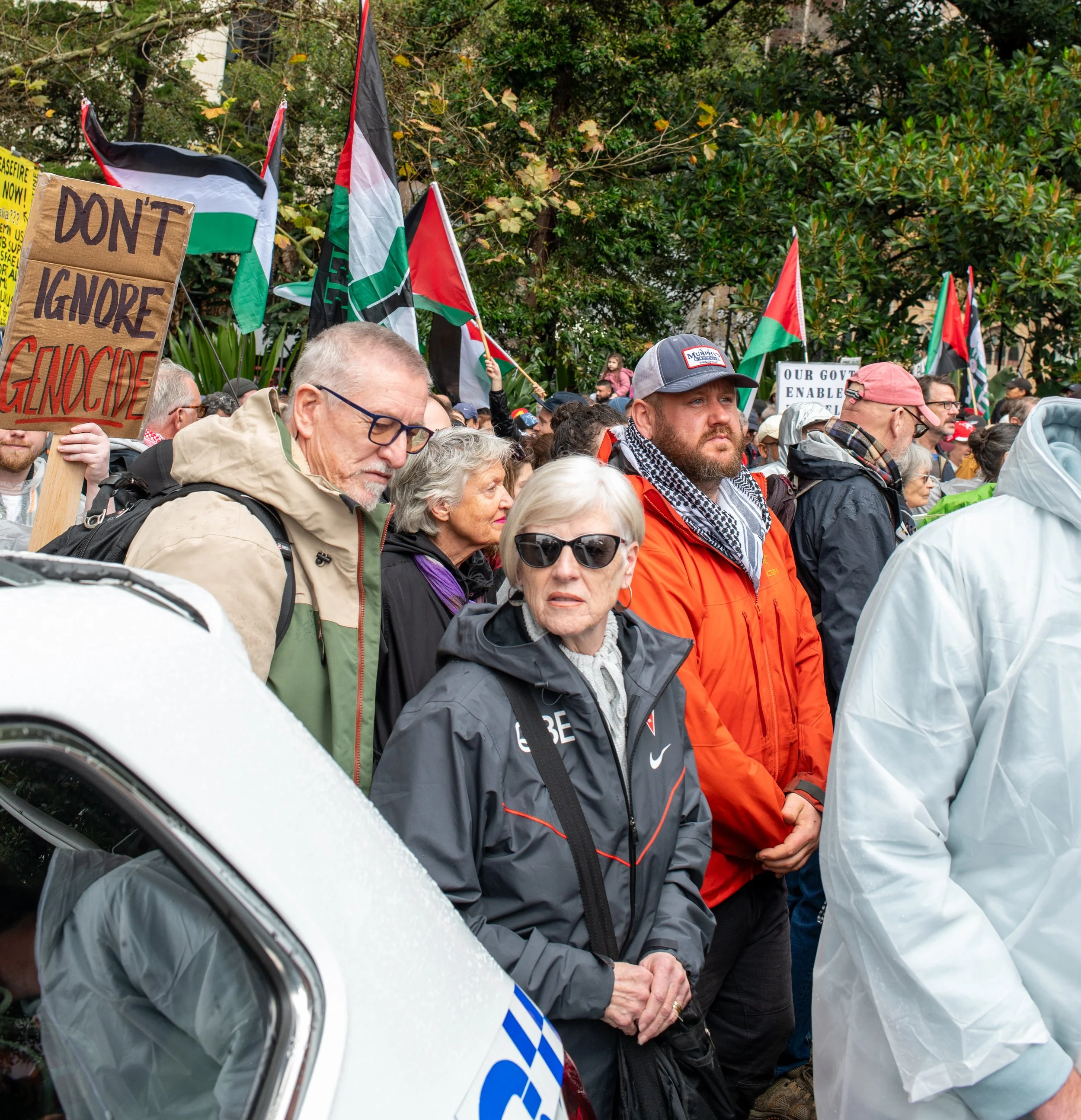 People participating in a protest or rally, holding flags and signs with messages like 'Don't Ignore Genocide,' surrounded by trees and wearing rain jackets and hats.