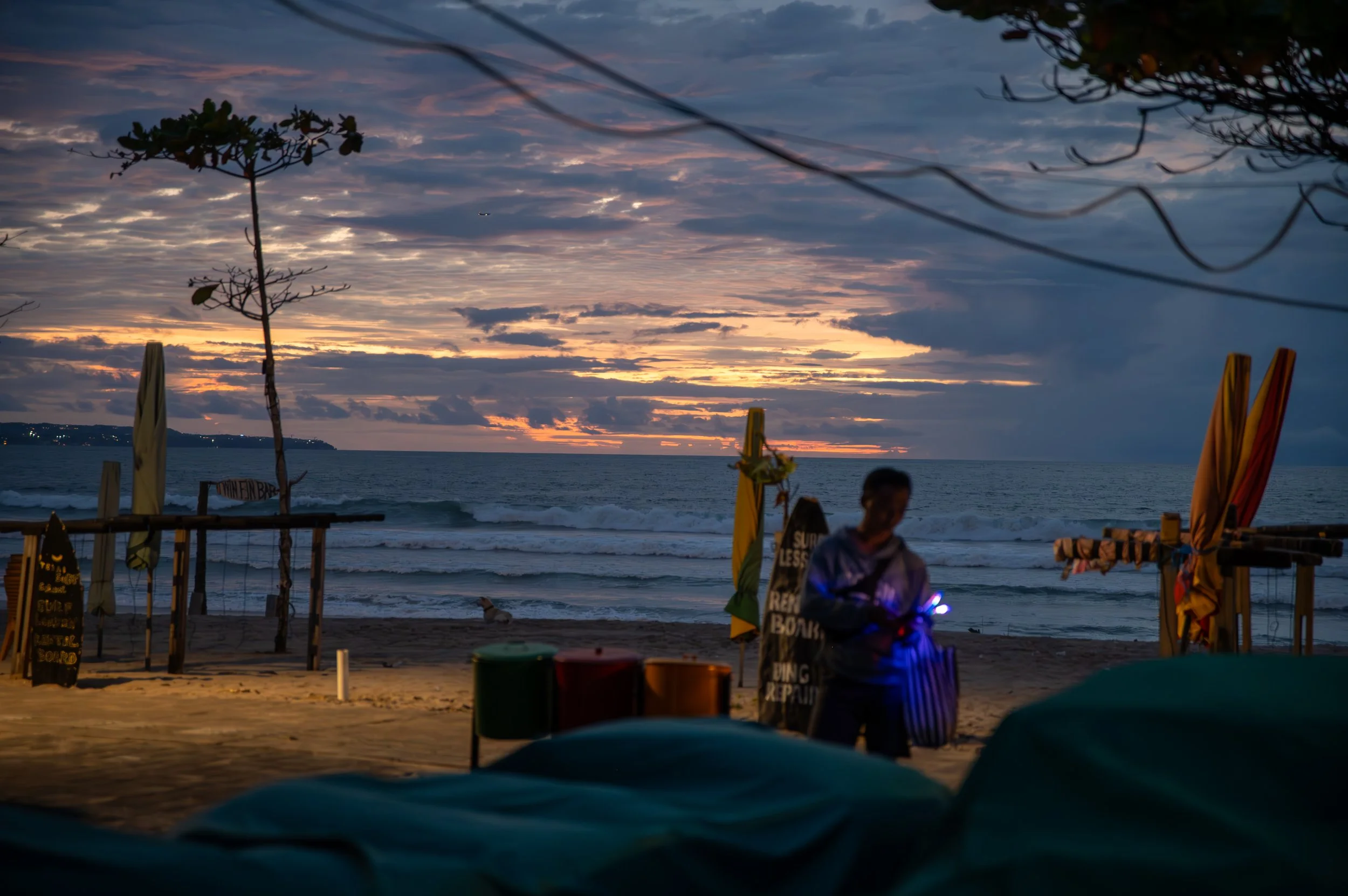 A sunset over the ocean with clouds in the sky, outlined by trees and beach chairs, with a person in the foreground wearing a hoodie and illuminated by small lights.
