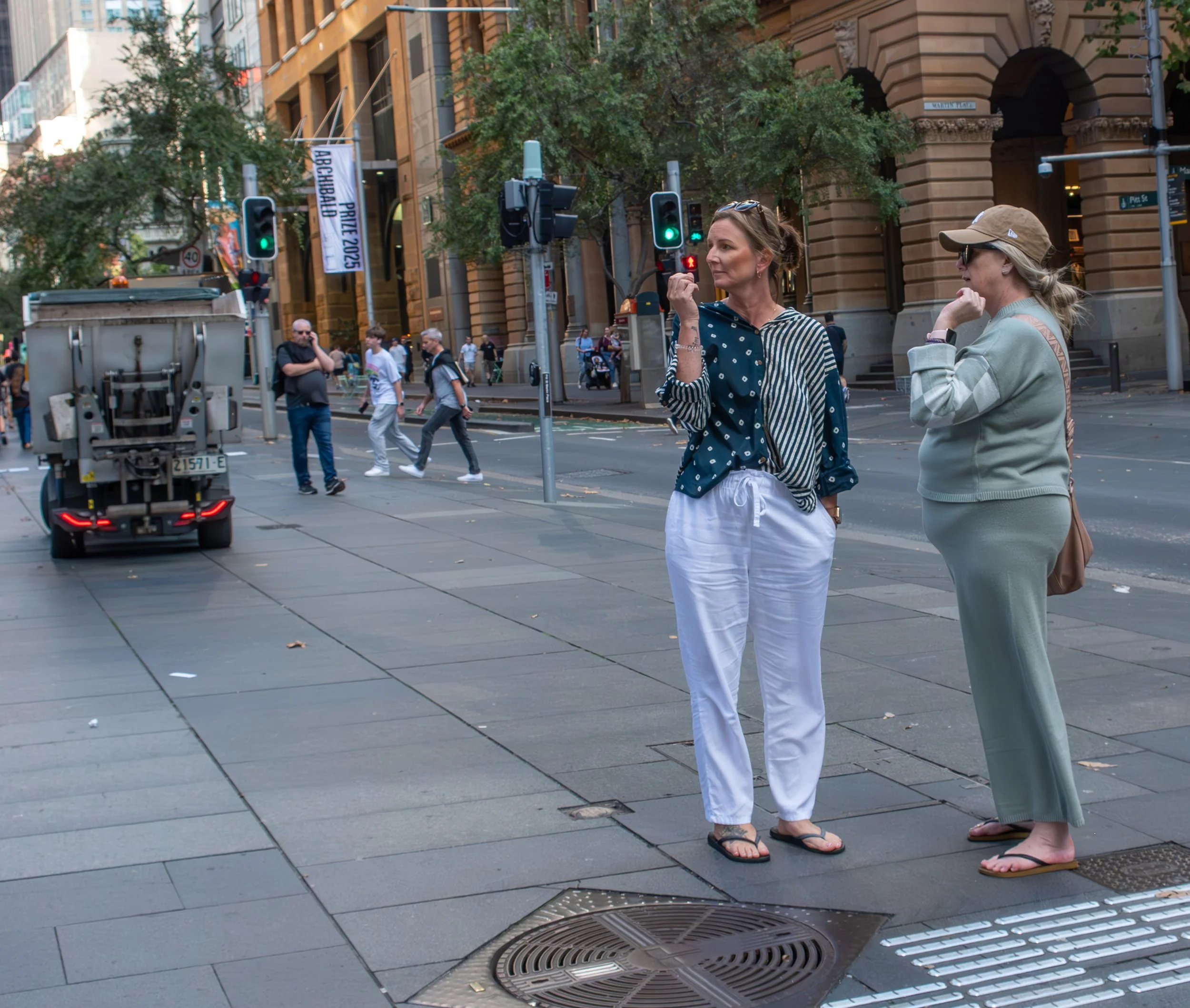 Two women standing on a city sidewalk near a crosswalk, with one woman wearing a blue patterned shirt and white pants, and the other in a beige tracksuit, both wearing flip-flops and sunglasses. People walk across the street in the background, along with traffic signals and buildings.