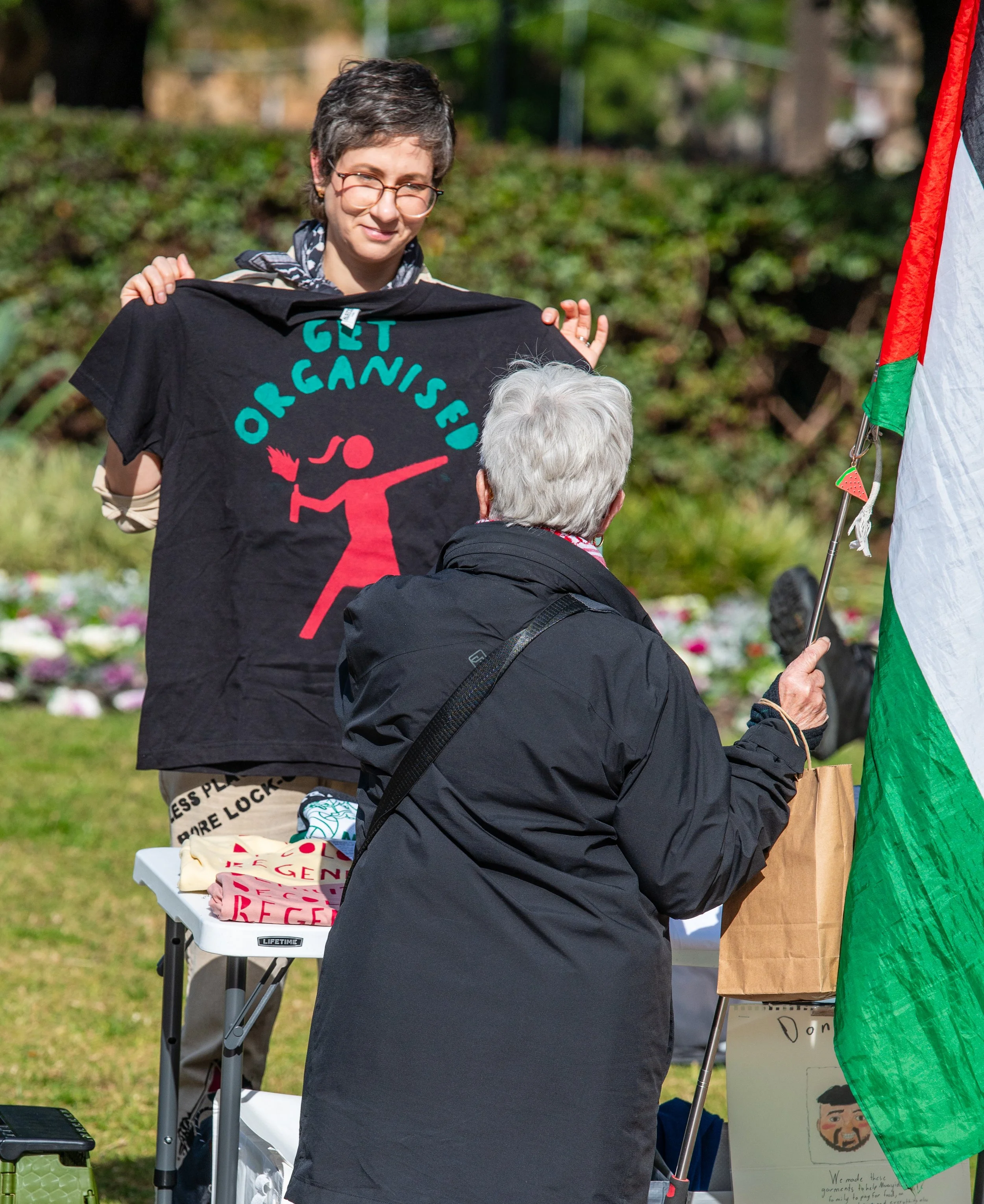 A young woman holding a black T-shirt with message 'Get Organised' and a figure illustration, standing outdoors behind a table, talking to an older woman with white hair who is holding a flag. The scene appears to be at a protest or rally.