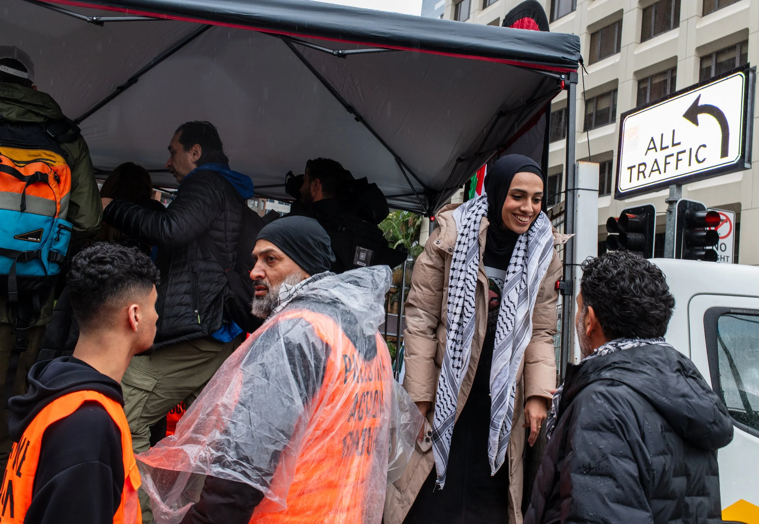 Group of people, including a woman with a headscarf, interact under a canopy on a rainy day in an urban setting with traffic signs visible in the background.