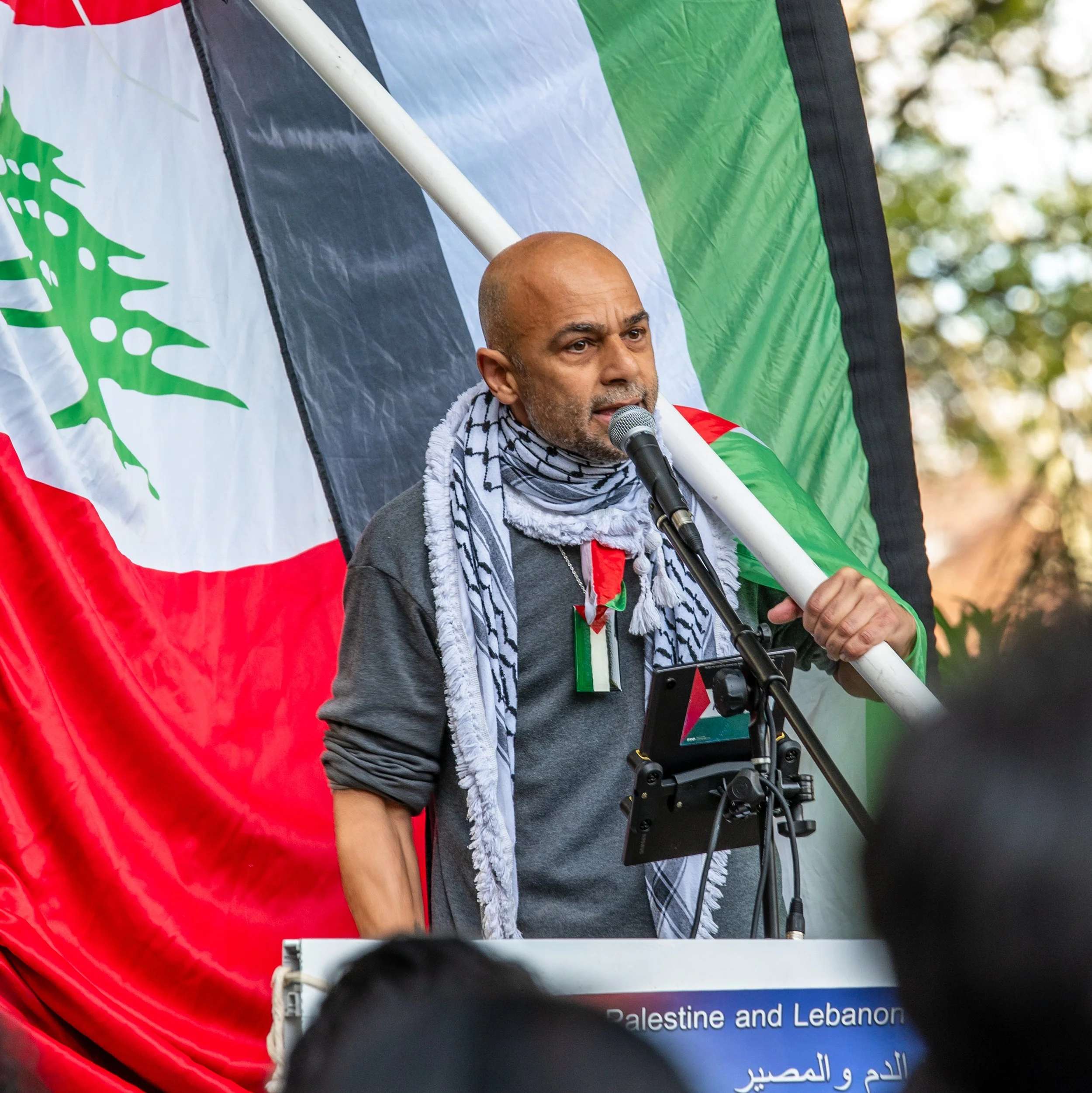 A man speaking into a microphone during a rally, holding a large Palestinian flag with a camouflage scarf around his neck, standing behind a sign that references Palestine and Lebanon.