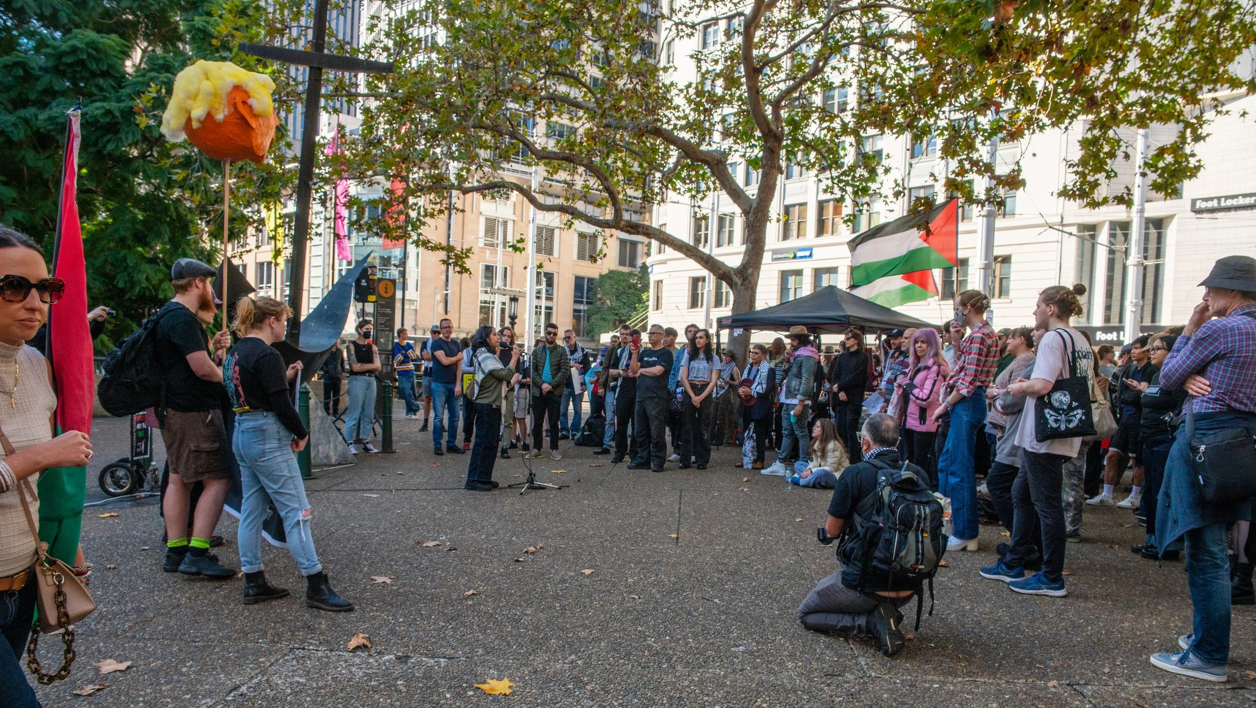 People gathered at a protest or rally in an urban park, with some holding Palestinian flags and a large puppet-shaped balloon of a face. Participants are standing or sitting on the ground, listening to a speaker at a microphone. There are trees, buildings, and tents in the background.