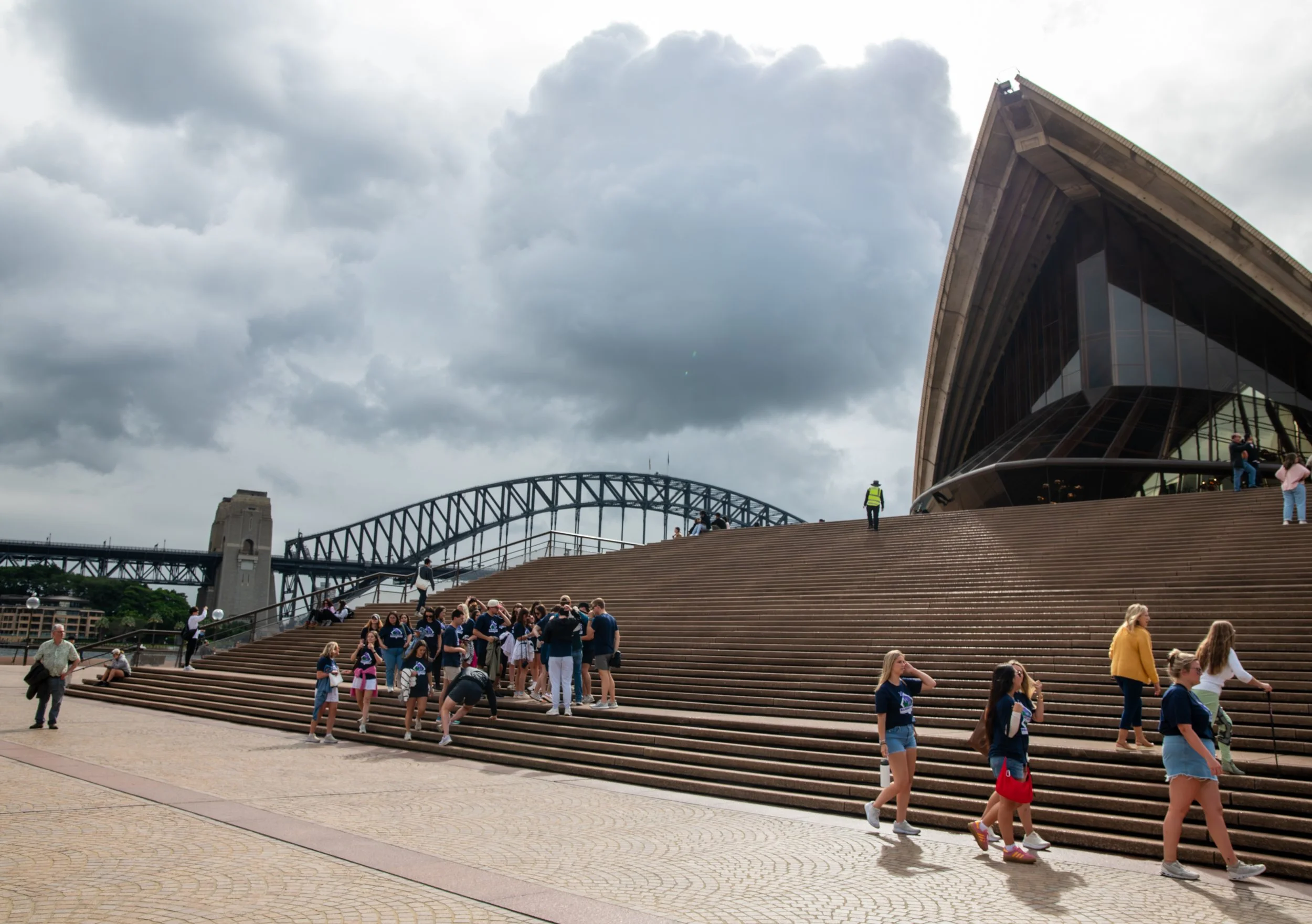 People gather on the large, stepped wooden stairs outside the Sydney Opera House with the Sydney Harbour Bridge in the background under cloudy skies.