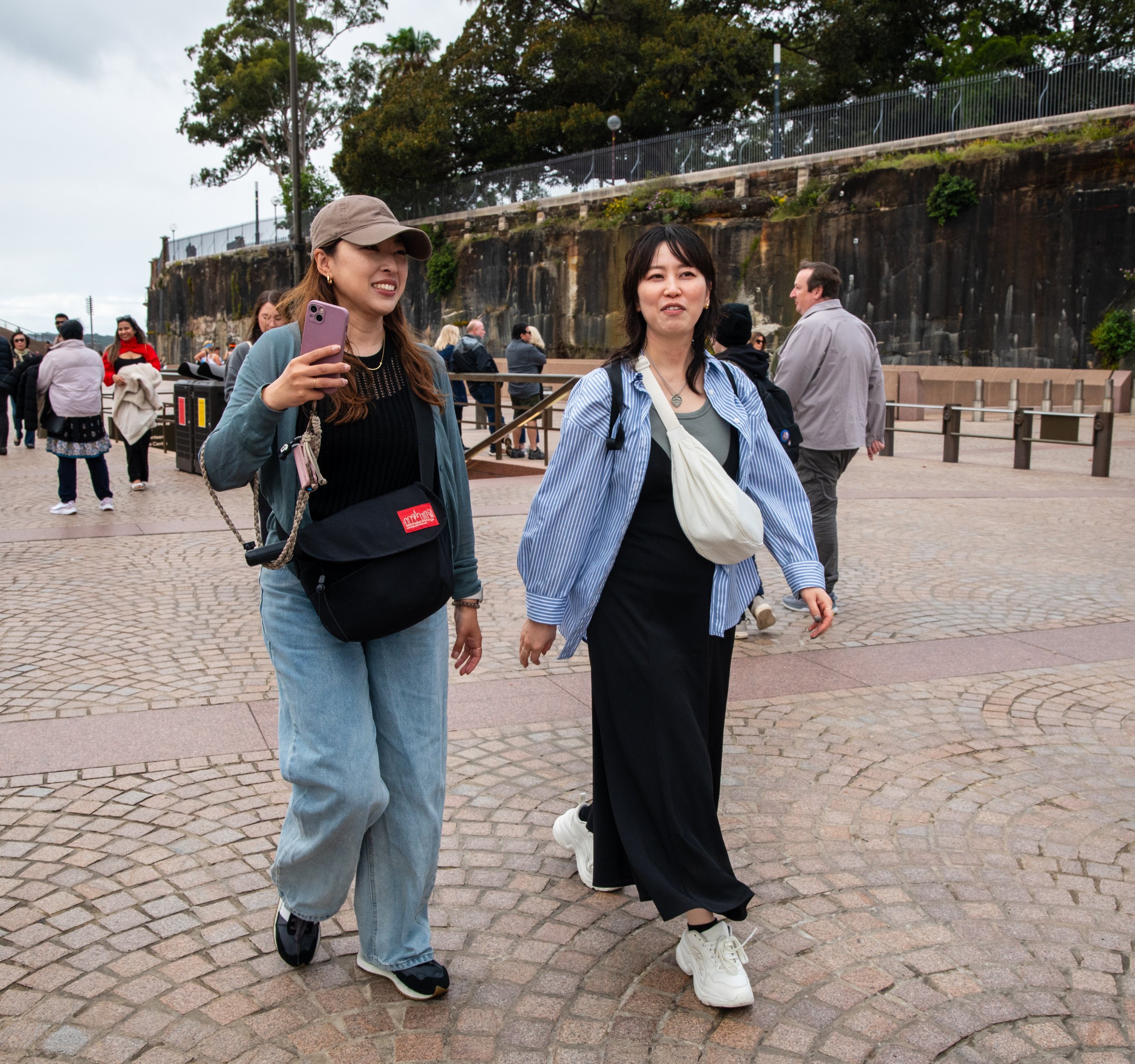 Two women walking on cobblestone pathway along a metal railing with rocks and green trees in background, surrounded by other people at an outdoor location.