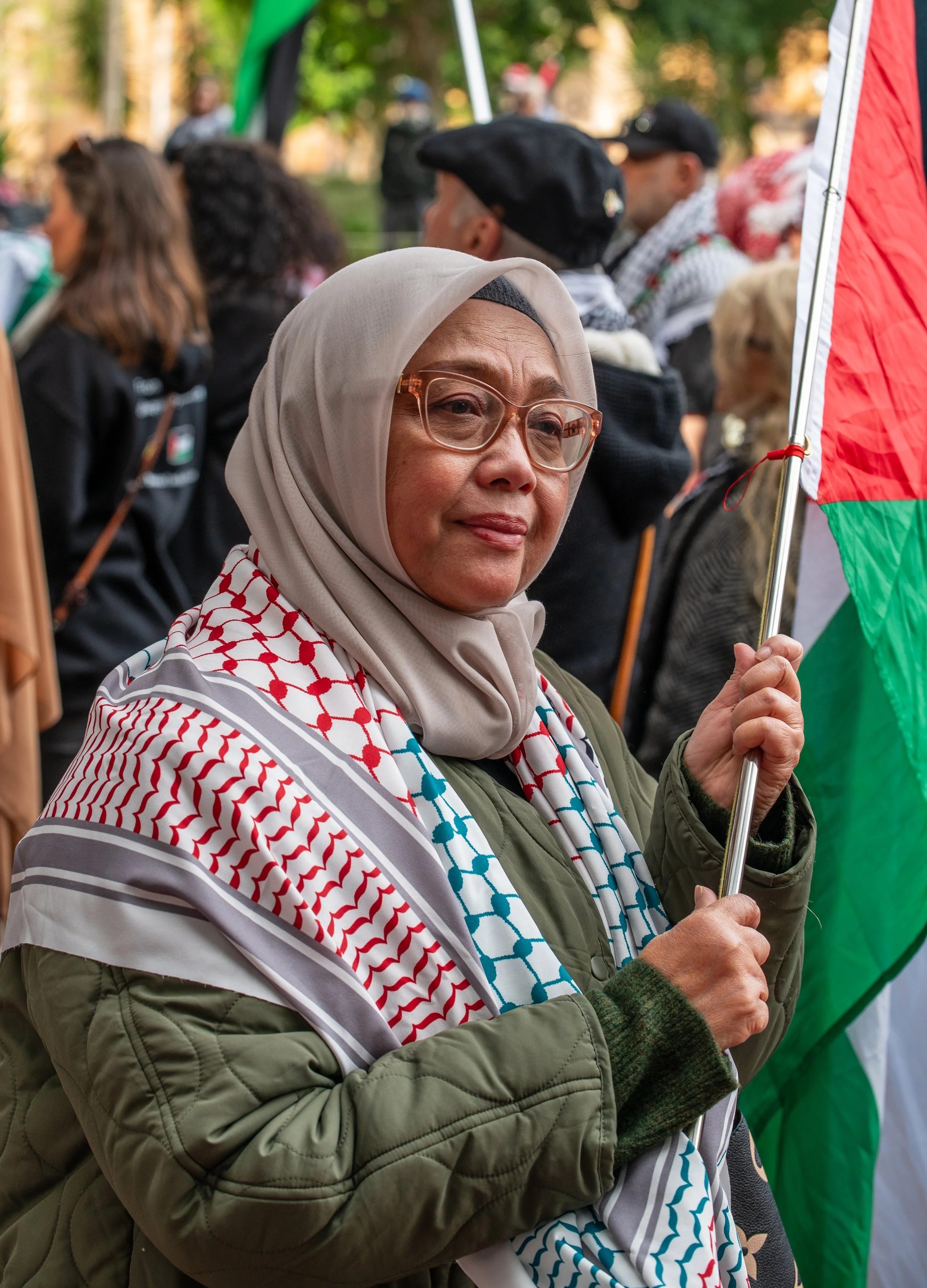 A woman wearing a beige hijab and glasses holding a Palestinian flag at a gathering or protest with other diverse individuals in the background.