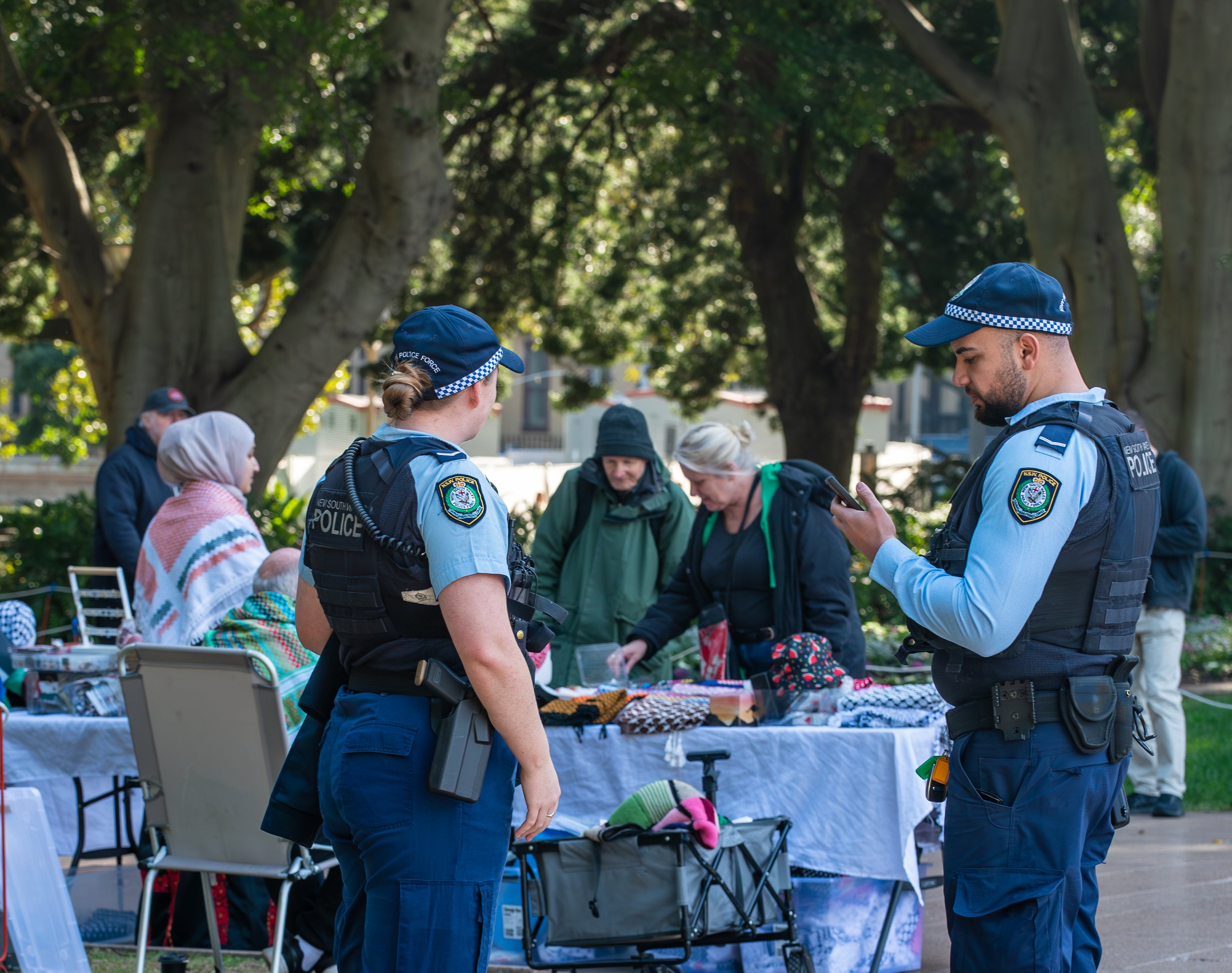 Two police officers, a man and a woman, stand near a table at an outdoor market or event, talking to shoppers. The officer on the right is looking at his phone, while the one on the left is facing the shoppers. Background includes people browsing items at a table, trees, and a sunny outdoor setting.