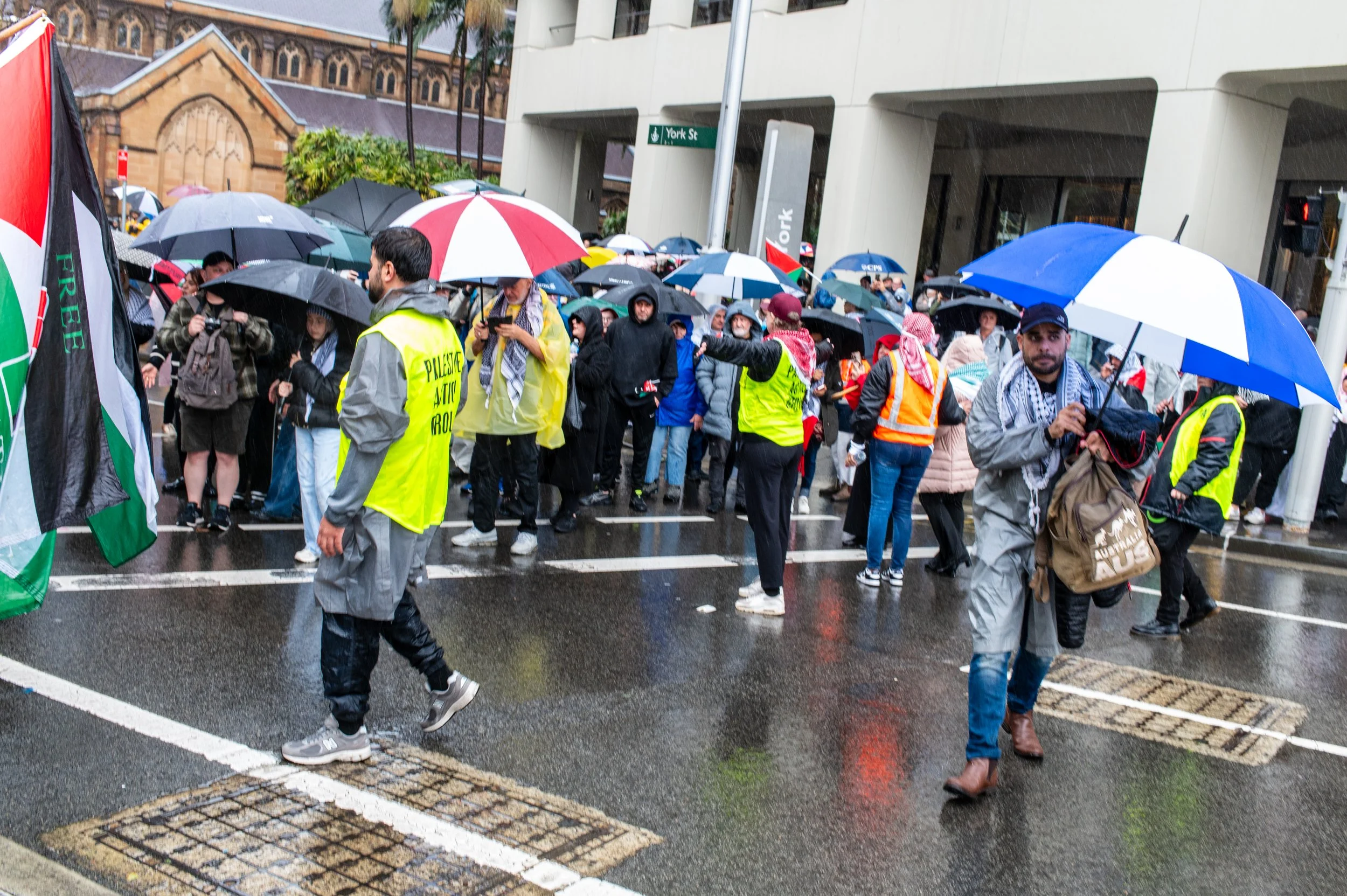 Crowd of people participating in a protest on a rainy day, holding umbrellas and some wearing high-visibility vests, on a city street near a building with a sign that reads 'York St'.