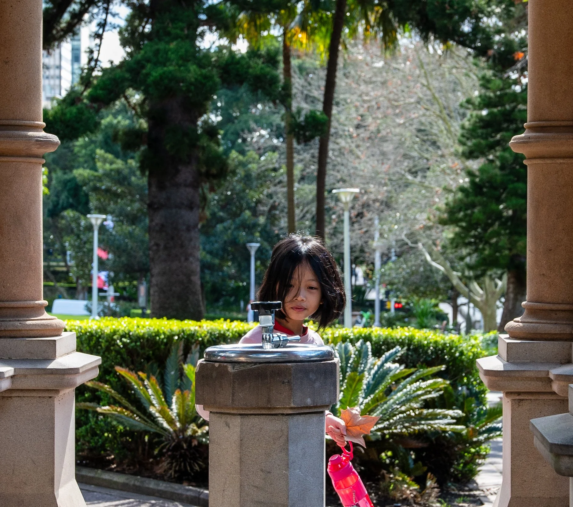 A young girl with shoulder-length black hair, holding a brown autumn leaf and a red water bottle, stands between two stone columns of a park pavilion, with trees, shrubs, and park lamps in the background.