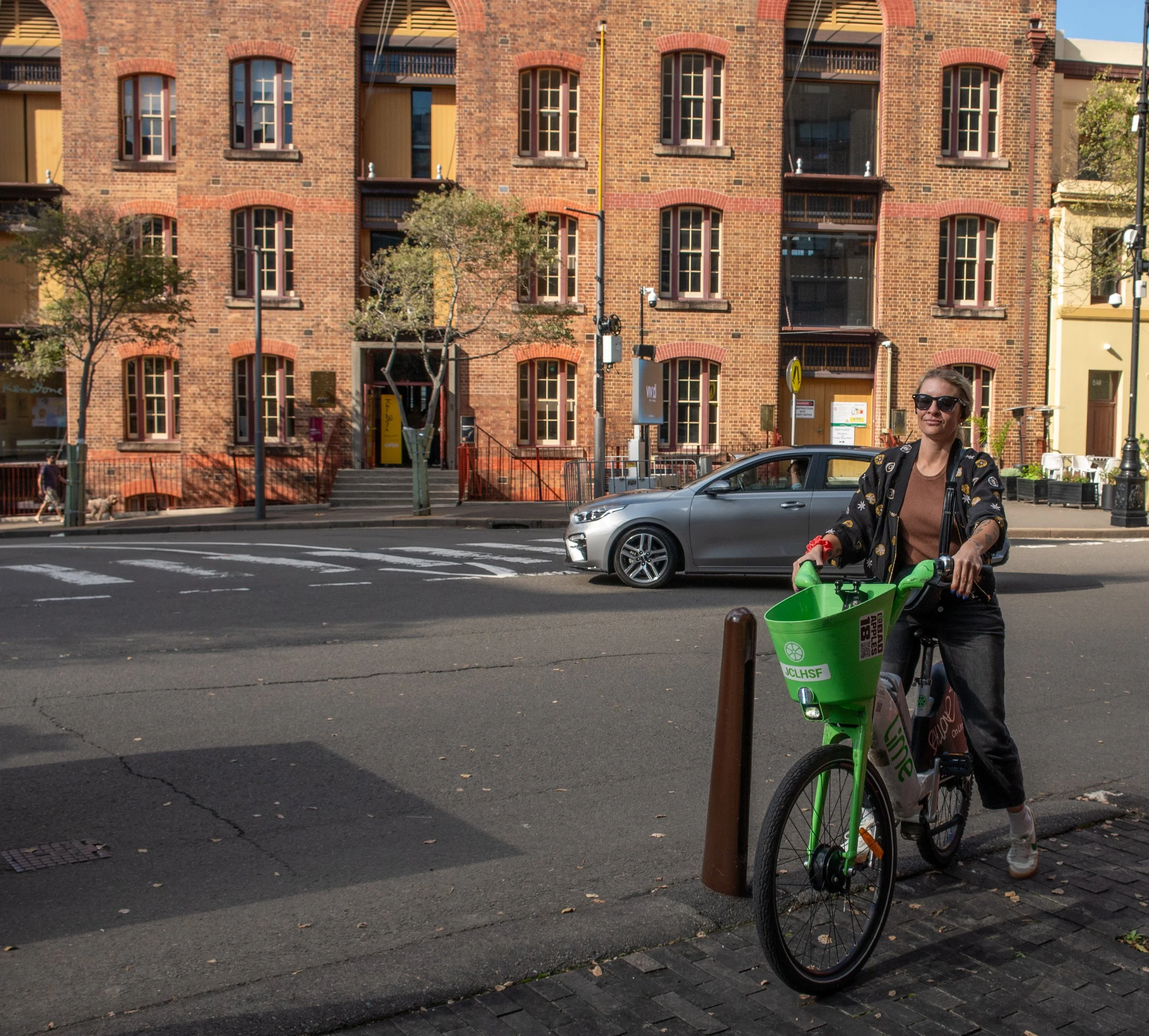 A woman stands next to a green CitiBike on a city sidewalk, with brick buildings and a street with cars in the background.