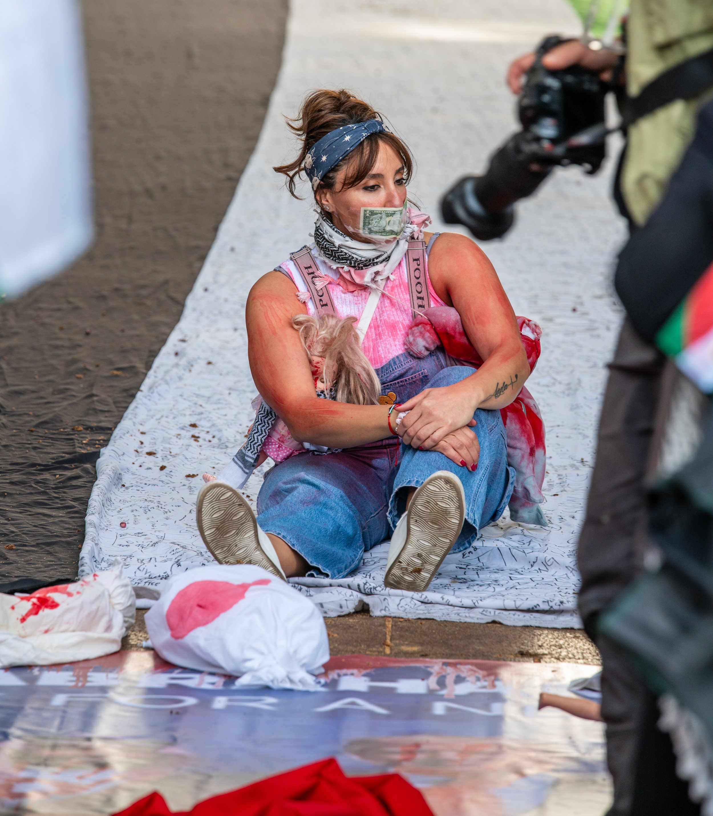 A woman sitting on the ground with a dollar bill on her face at a protest, surrounded by signs and other protest materials.